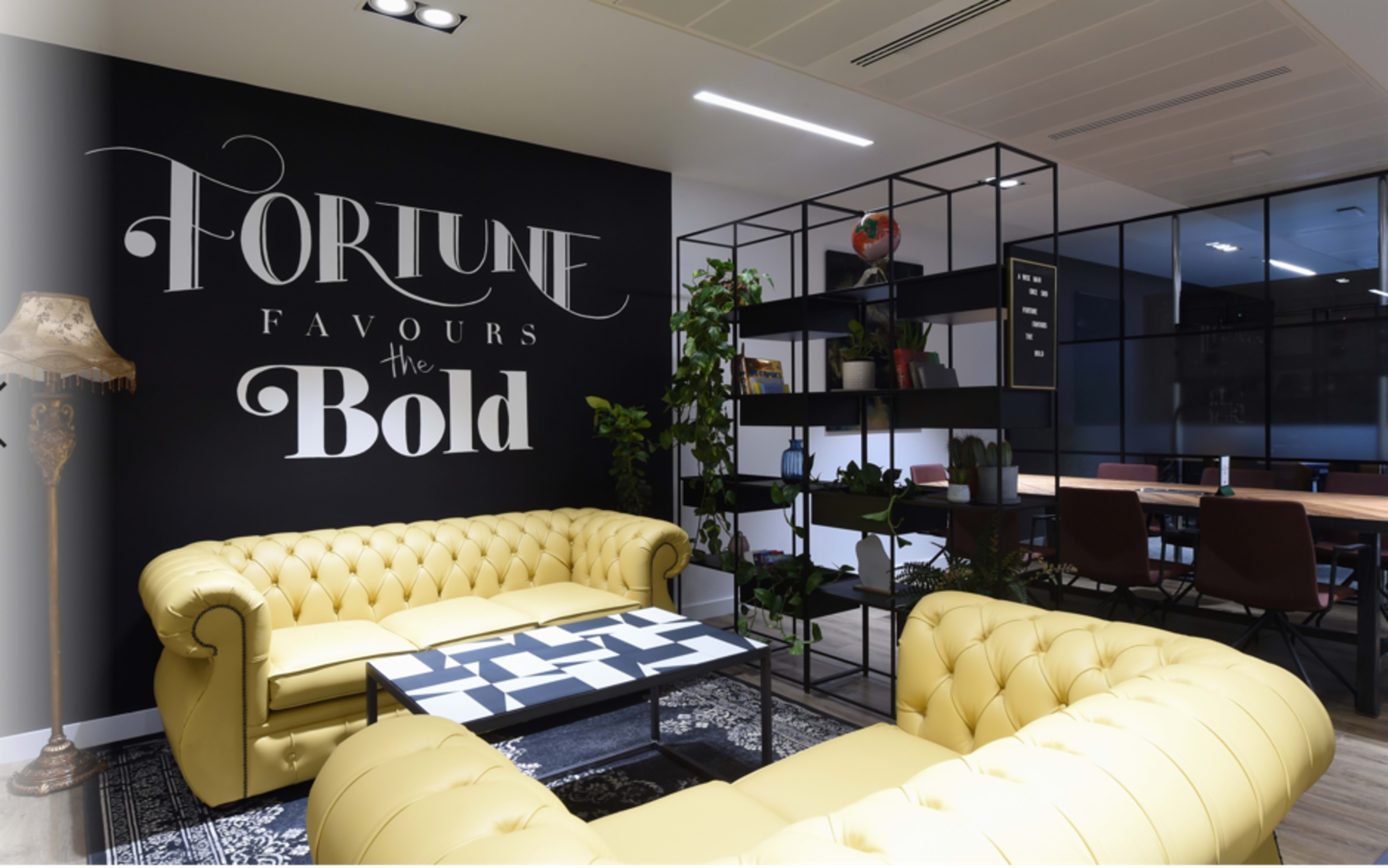 A modern office lounge area featuring yellow chesterfield sofas, a black and white patterned coffee table, and a wall with the phrase "Fortune Favours the Bold" in stylish lettering.