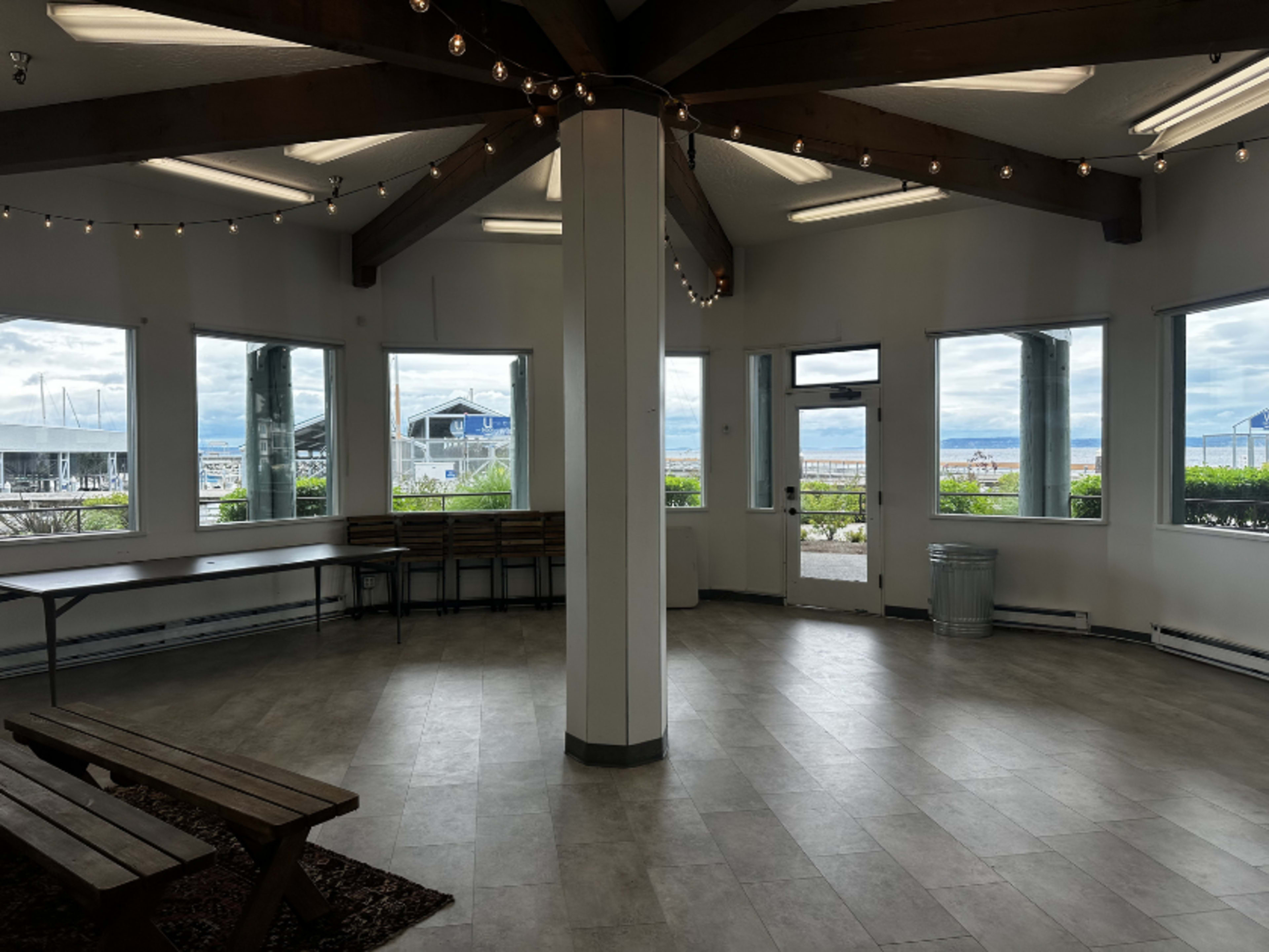 The image shows a spacious, well-lit interior room with large windows, a central column, and several wooden benches along the walls.