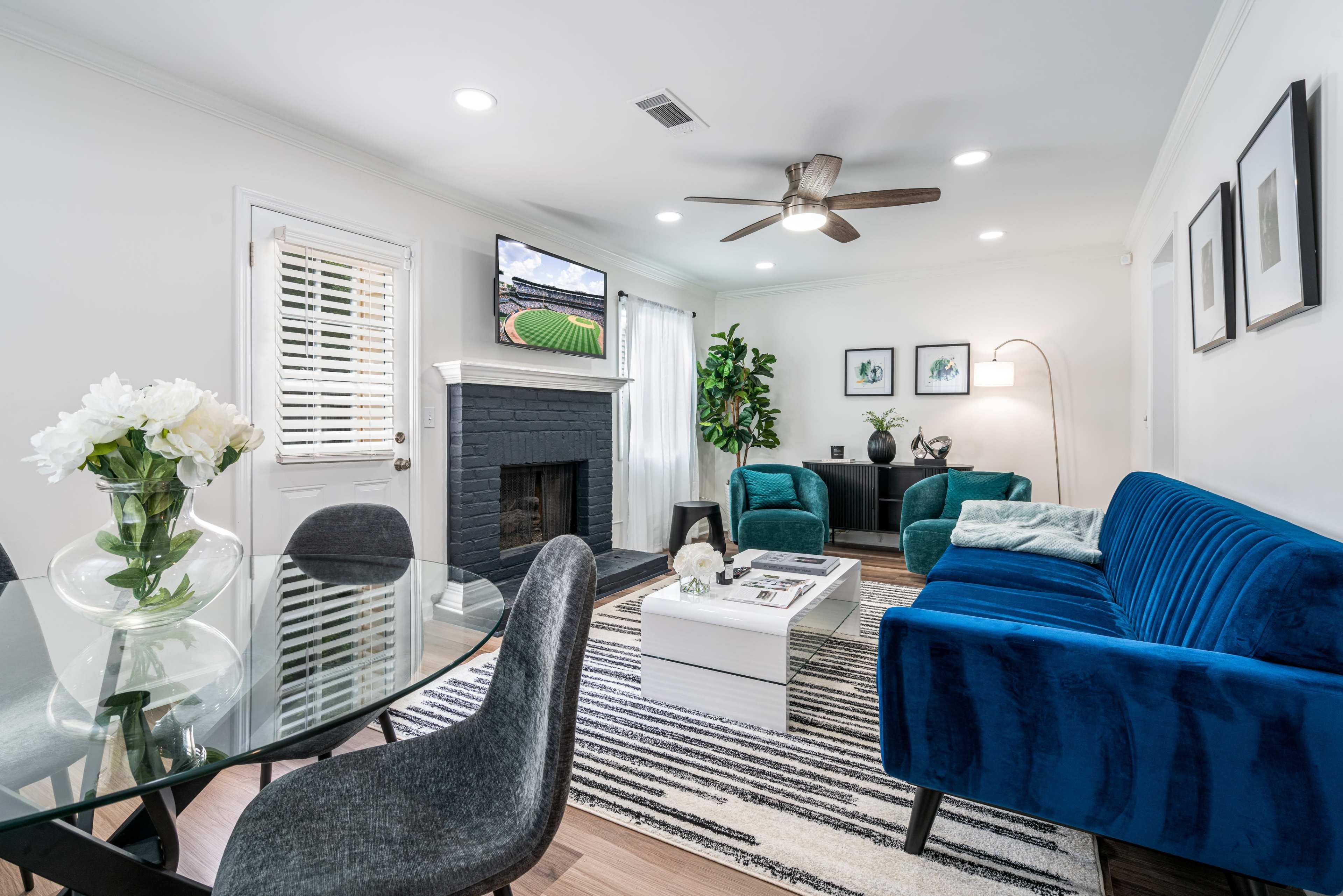 A modern living room with a blue velvet sofa, a glass dining table with floral decor, and a wall-mounted television above a black fireplace.