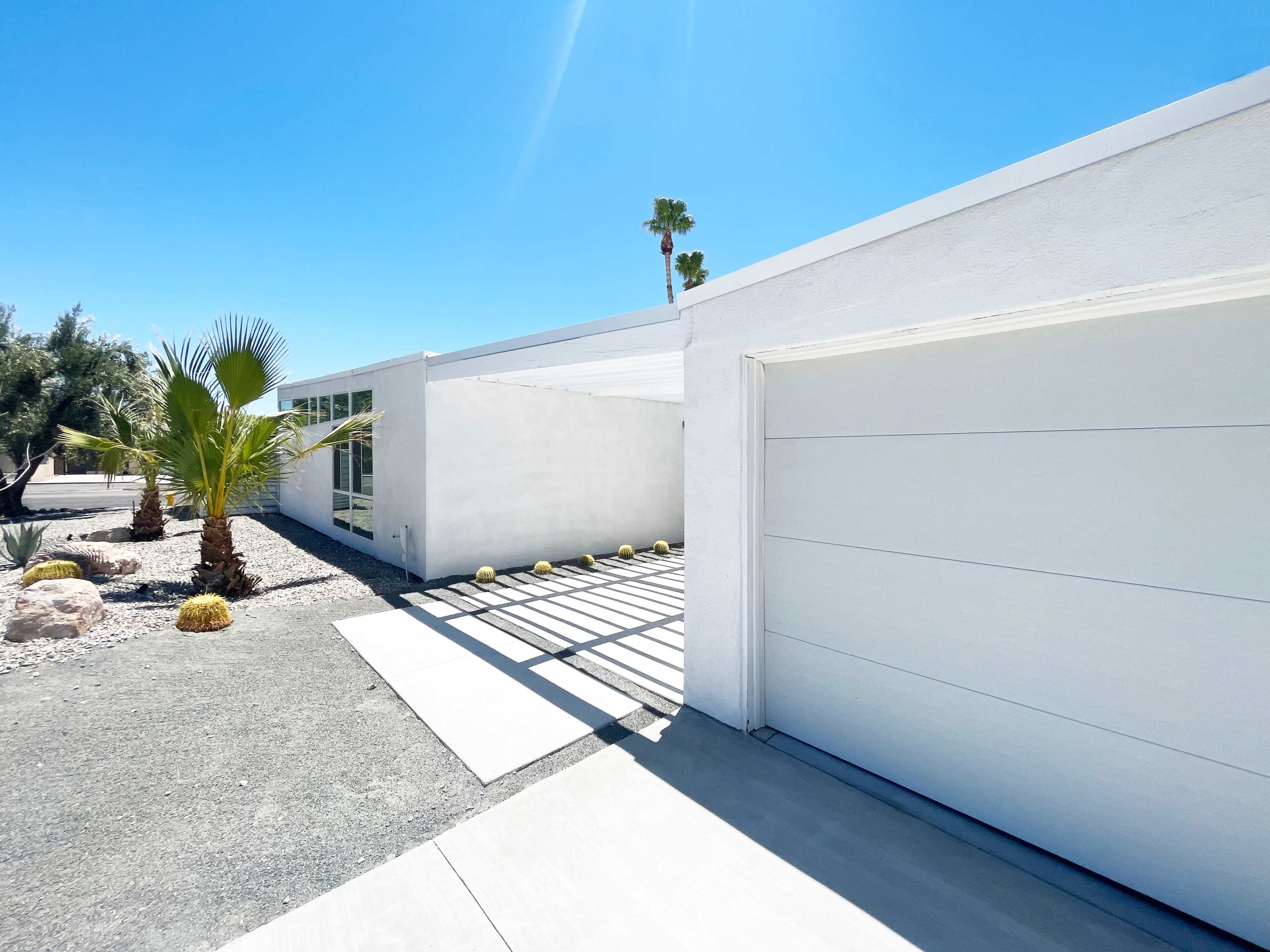 A modern white house with a garage is surrounded by palm trees and pebbled landscaping under a clear blue sky.