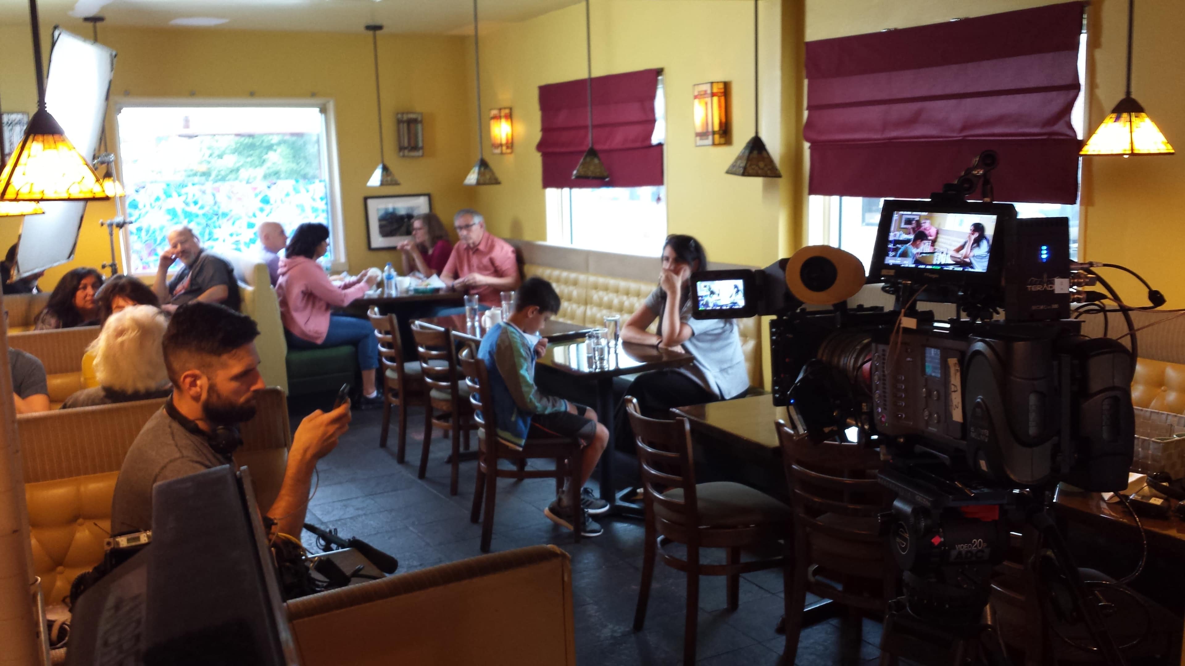 A film crew sets up cameras and equipment in a brightly lit restaurant, where diners are seated at various tables.
