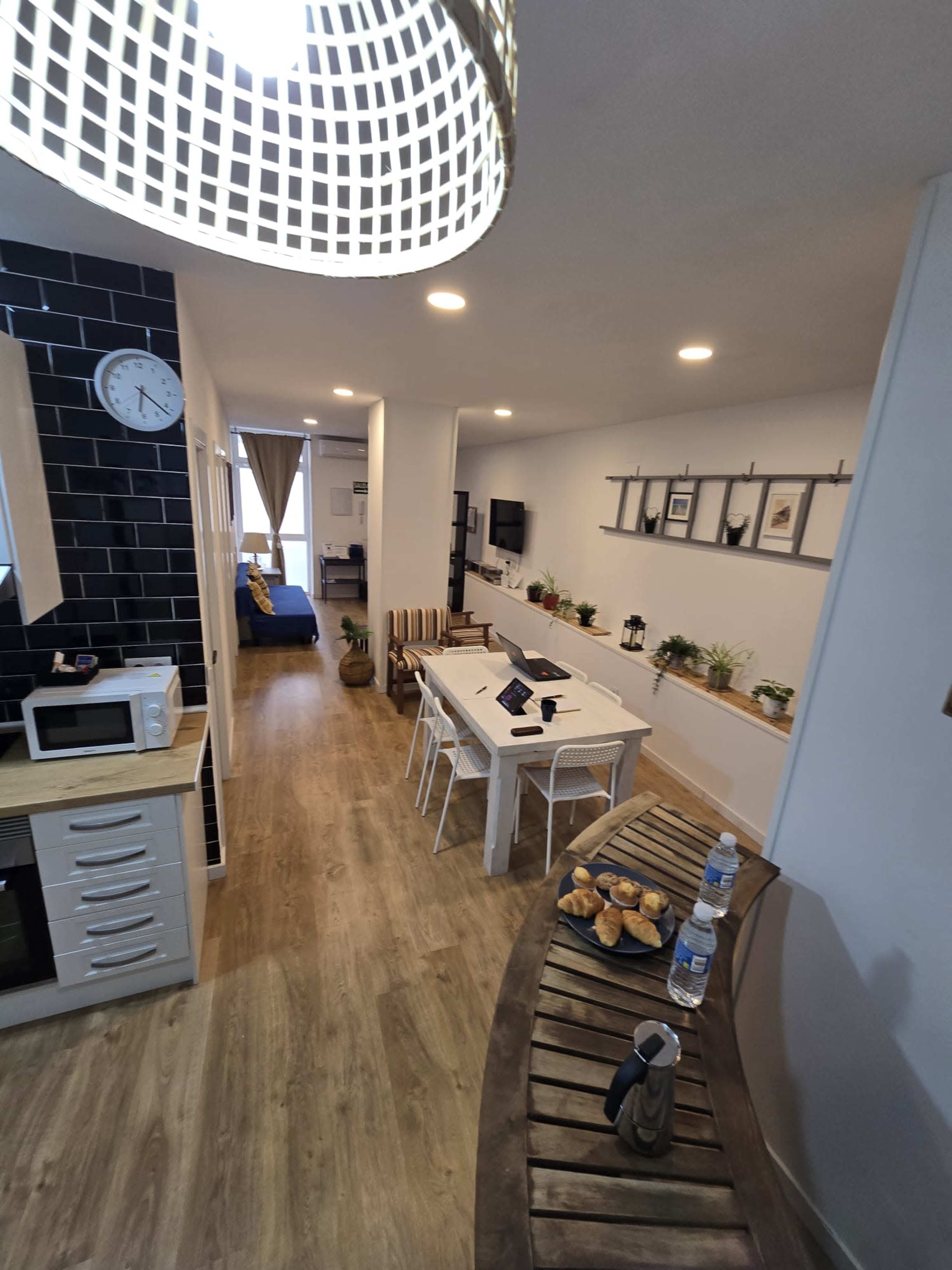 A modern kitchen and dining area are visible, featuring a long white table, a wooden side table with beverages, and a view into a living room with a sofa and wall-mounted TV.