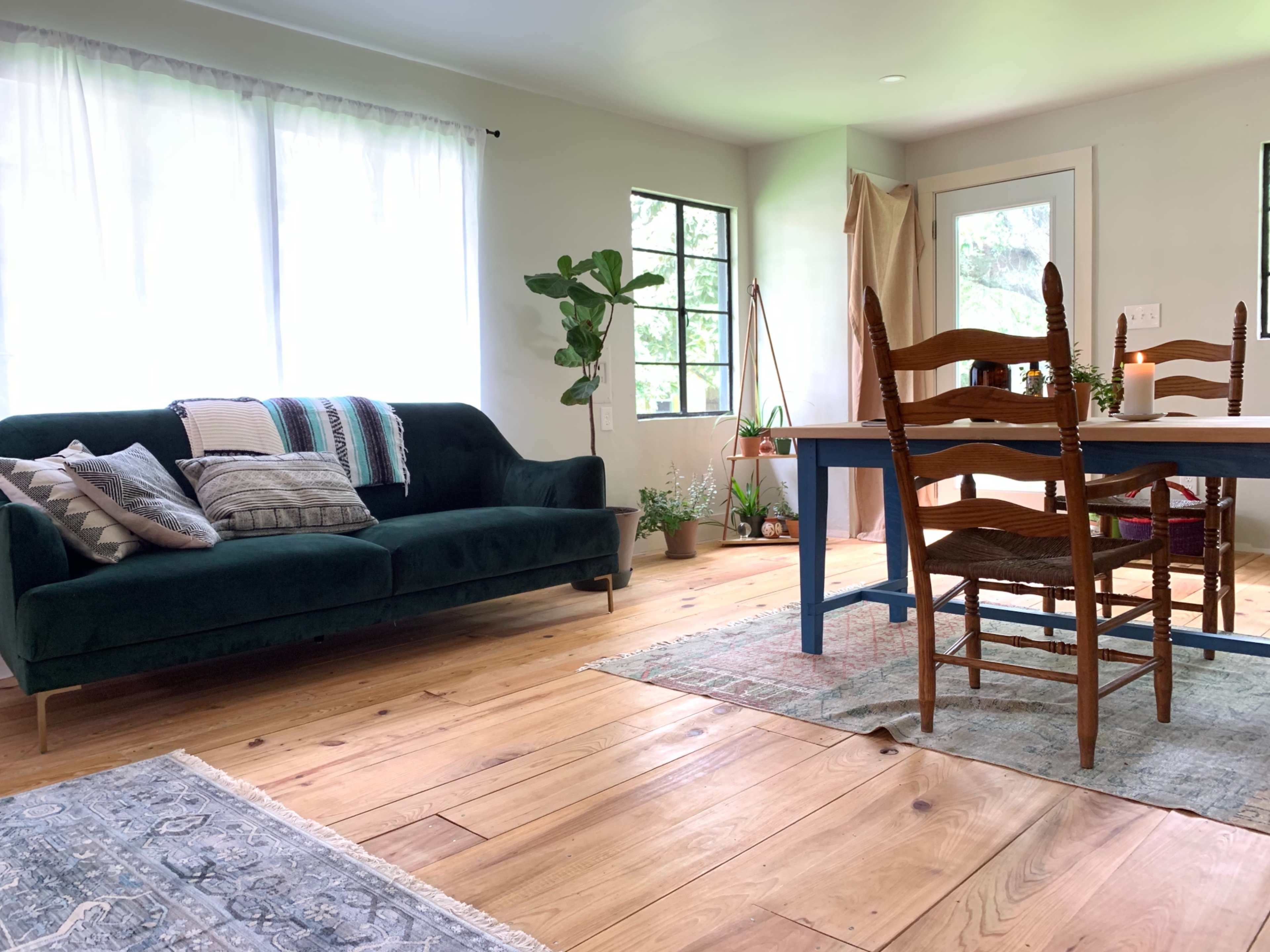 A living room with a green sofa, a wooden dining table with chairs, and large windows allowing natural light to enter.