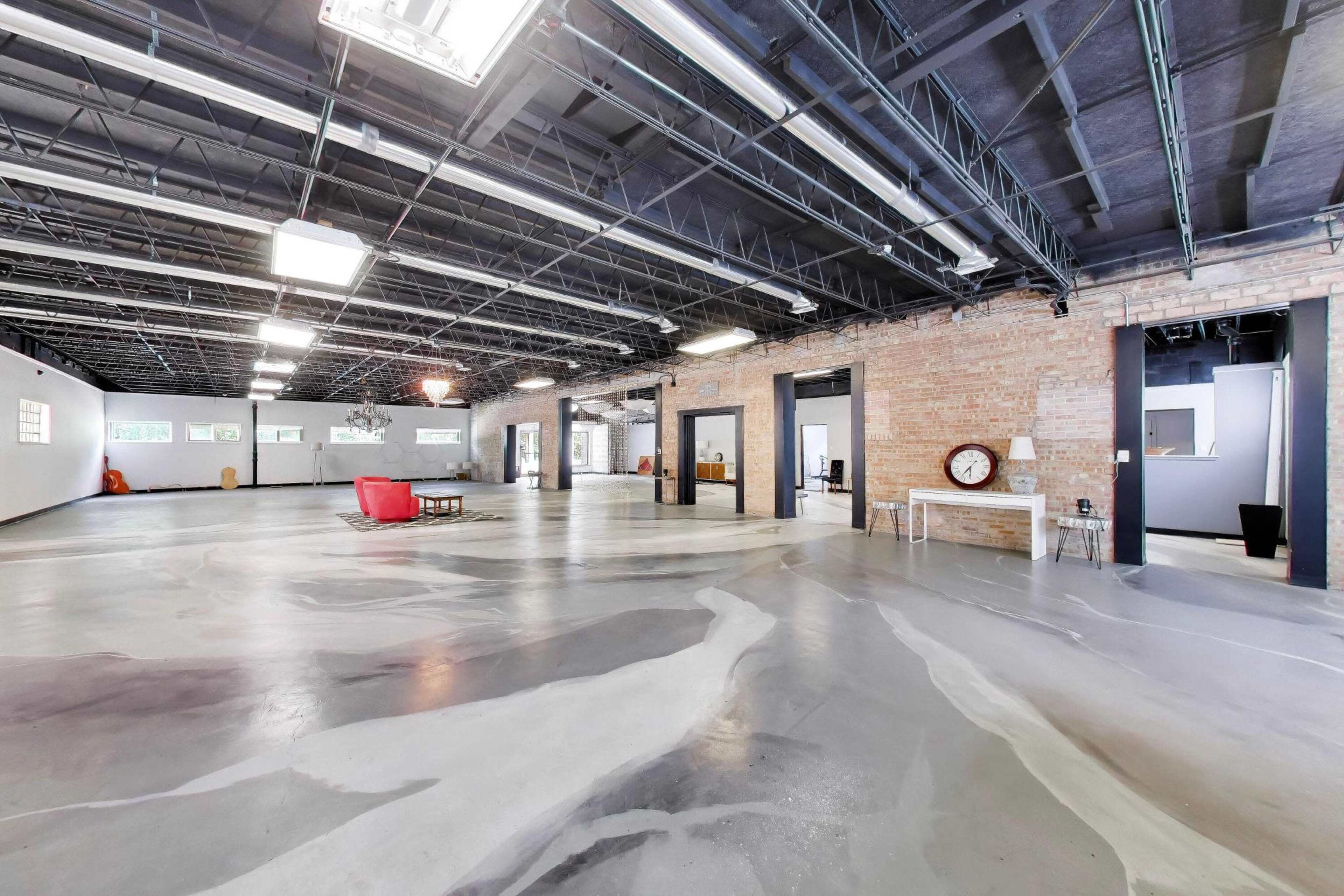 The image shows a spacious, industrial-style interior featuring polished concrete floors, exposed brick walls, and large overhead lights.