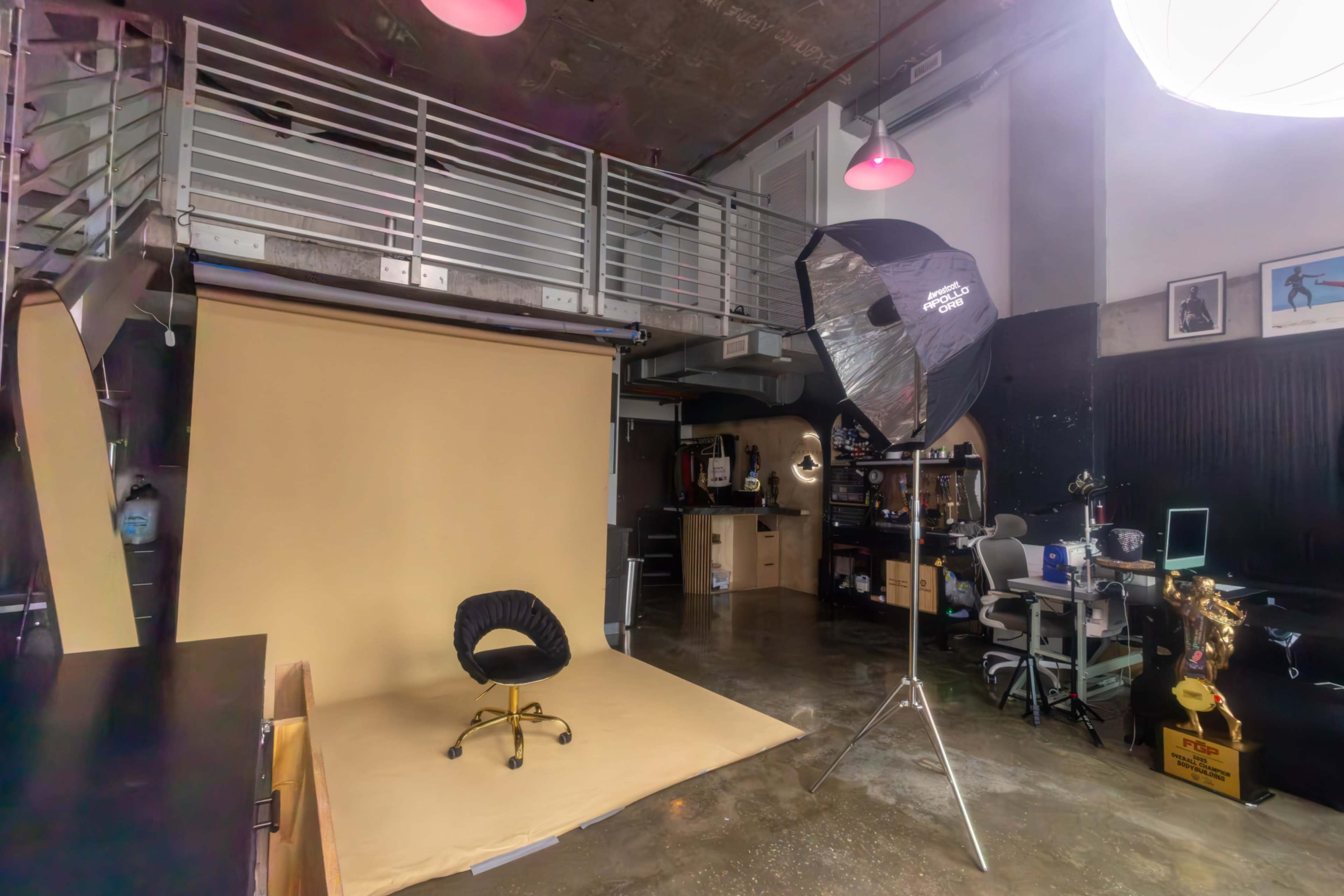 The image shows a photography studio with a beige backdrop, a black chair on wheels, and various photography equipment and setups in the background.