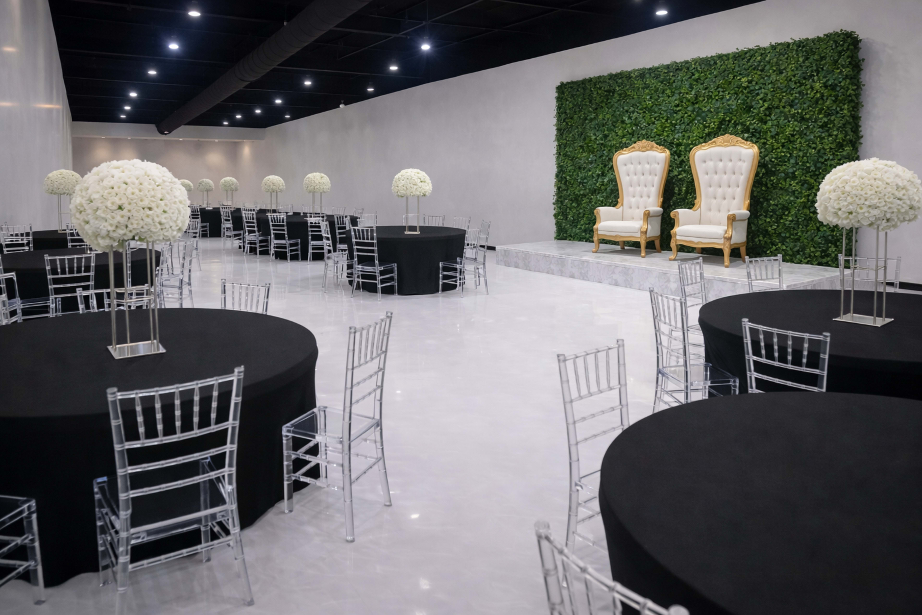 A spacious, elegantly decorated event hall features black round tables with clear Chiavari chairs and a pair of golden throne-like chairs against a green foliage backdrop.