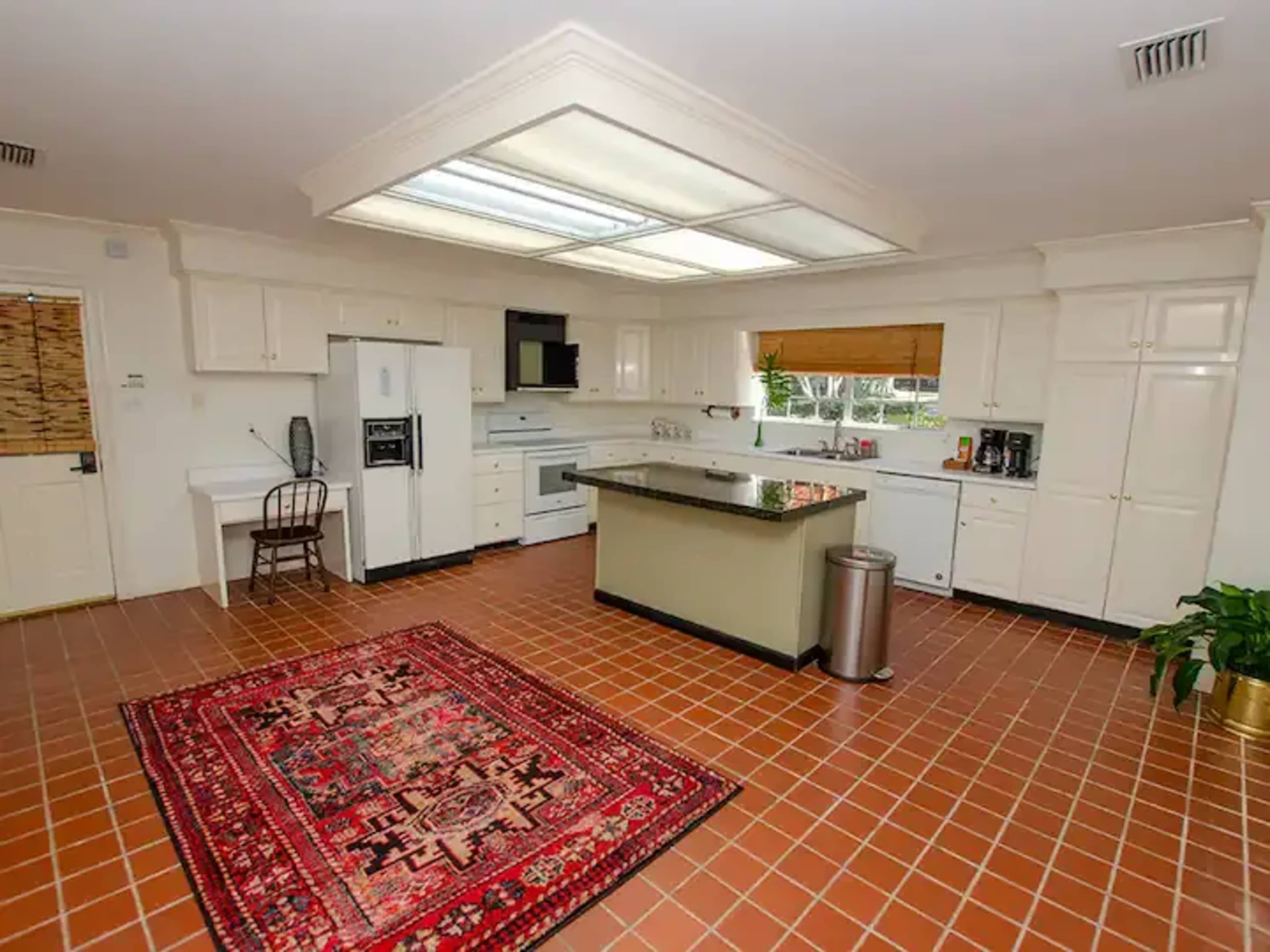 A spacious kitchen features white cabinets, a central island, and a large red area rug on a tiled floor.