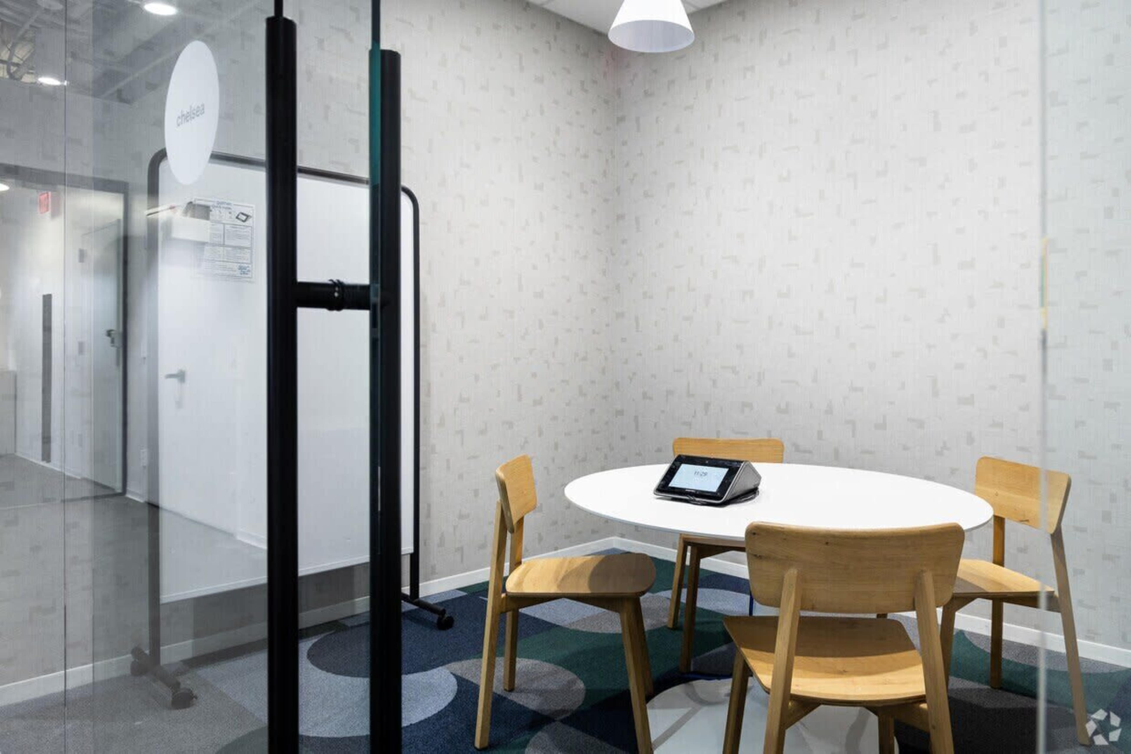 The image shows a small meeting room with a round white table, four wooden chairs, and a tablet display on the table.