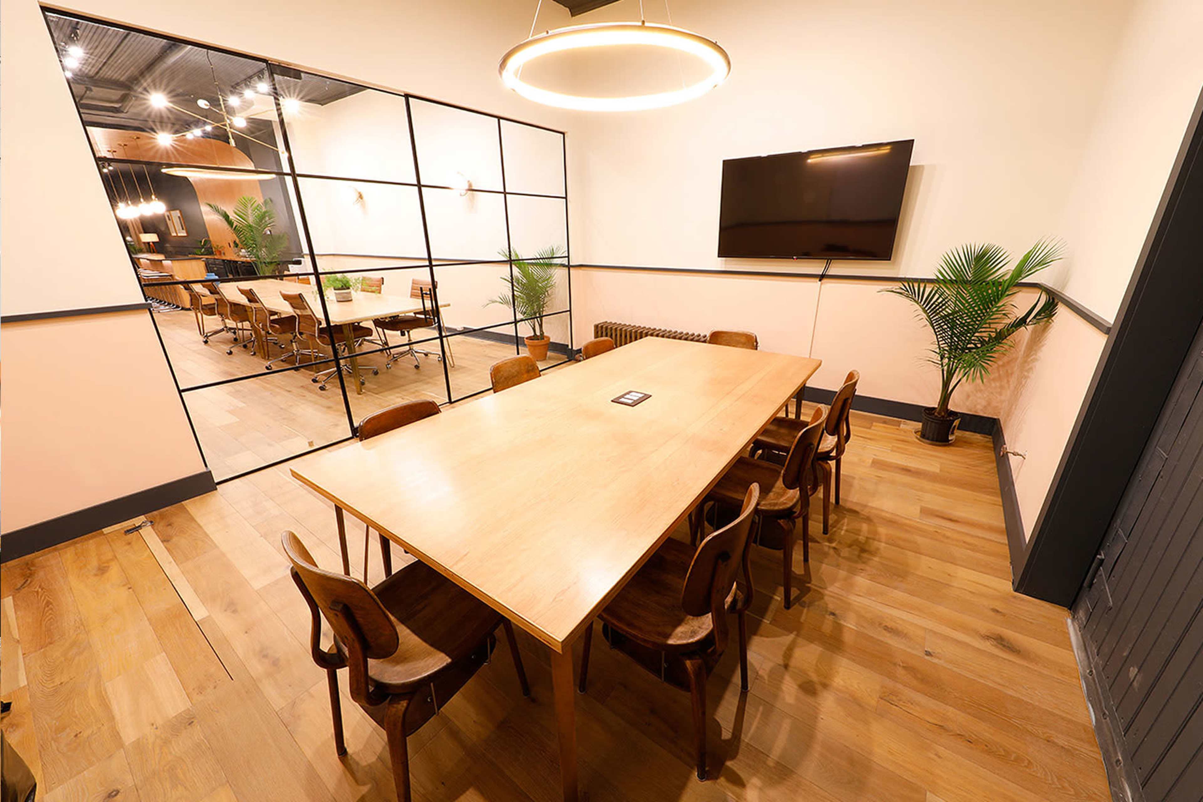 A spacious conference room features a large wooden table surrounded by eight chairs, with a wall-mounted TV and large windows leading to an adjacent room.