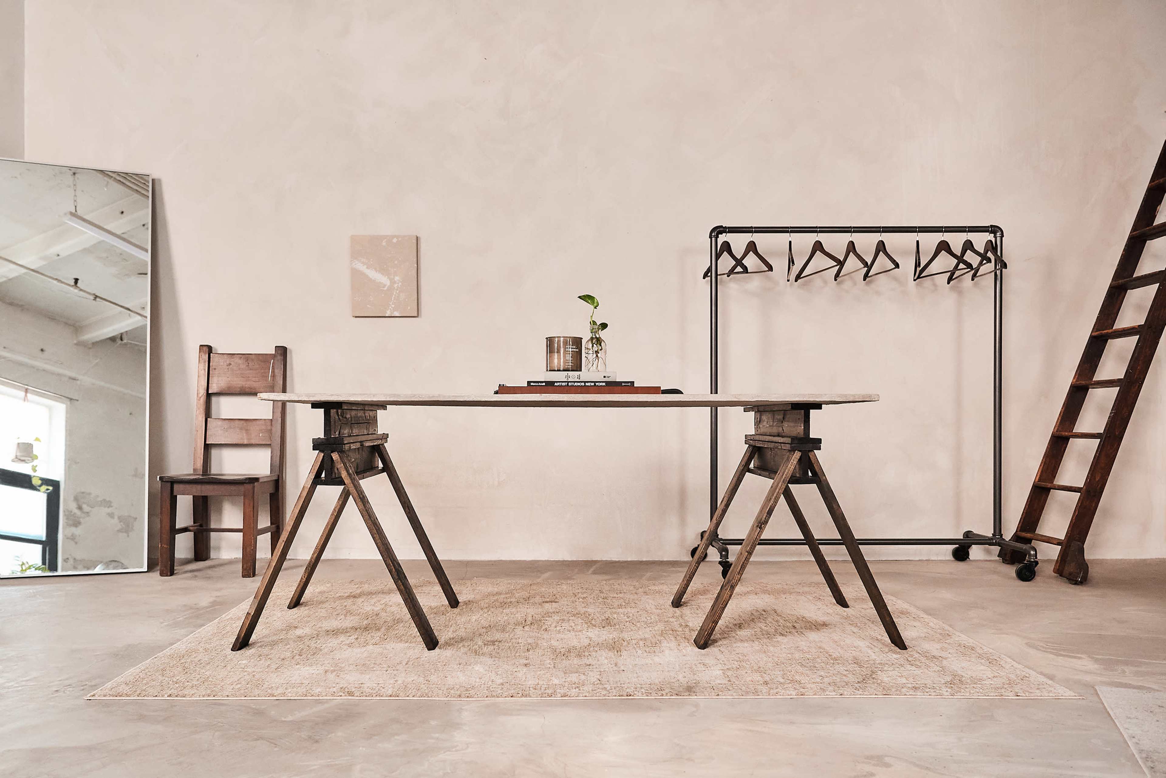 The image shows a minimalist room with a wooden table supported by metal sawhorse legs, alongside a chair, a clothing rack, and a leaning ladder against the wall.