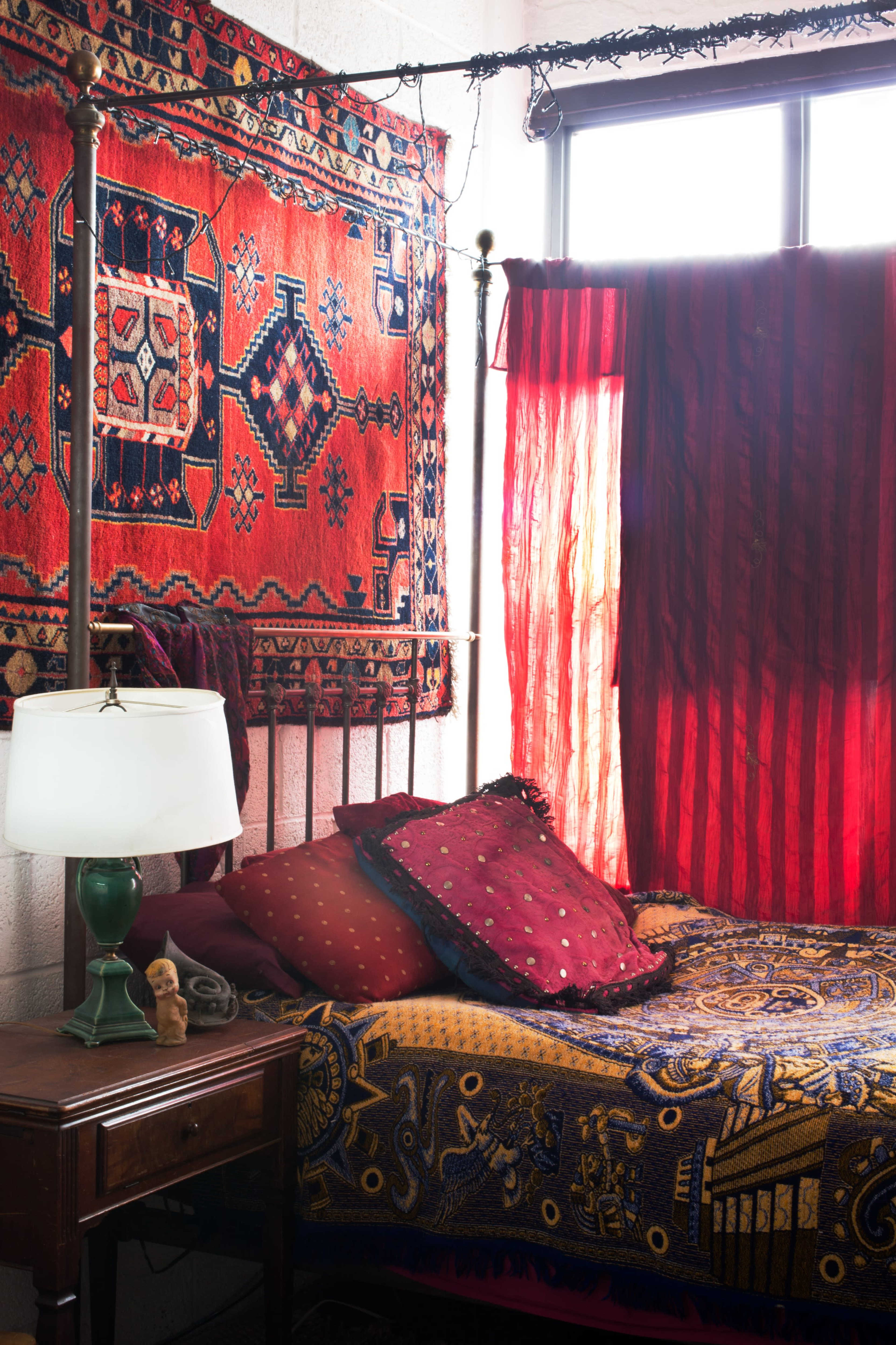 A bed with patterned bedding and pillows is positioned beside a table lamp and a wooden nightstand under a window draped with red curtains and adorned with a colorful tapestry.