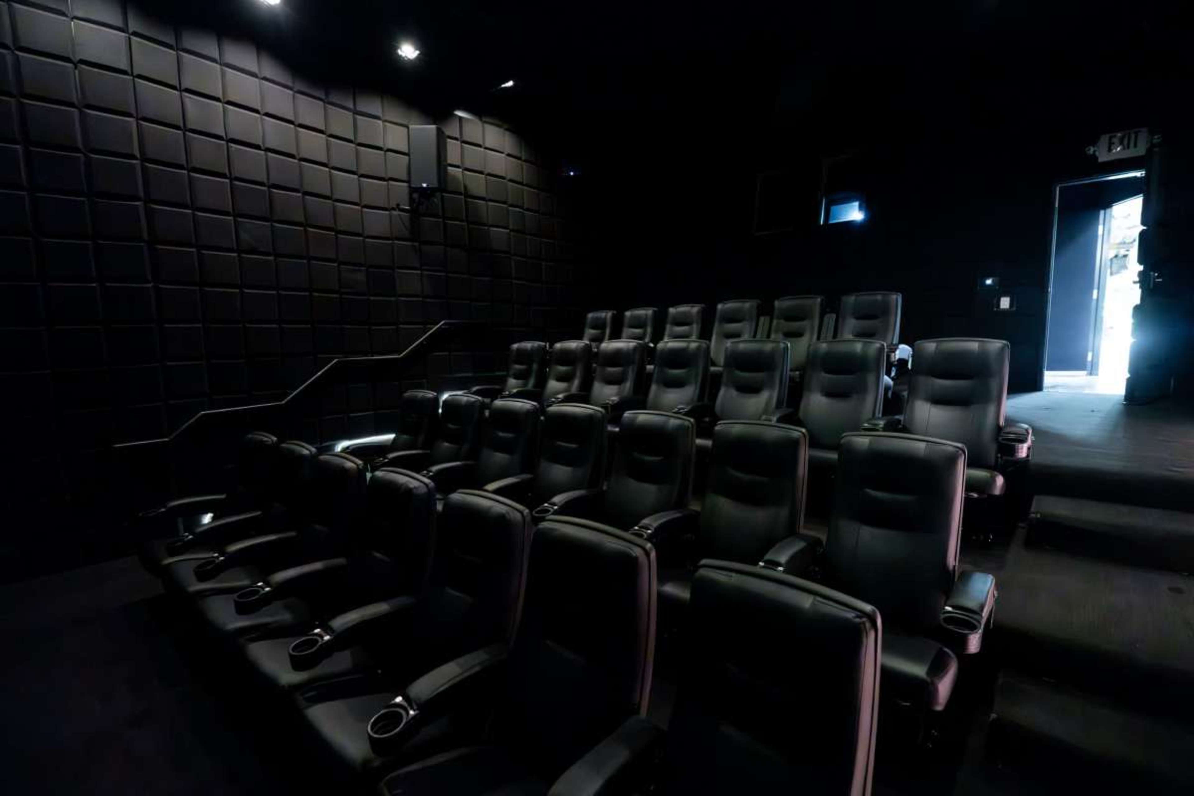 The image shows a dark, empty movie theater with rows of black reclining seats and a doorway illuminated by light.