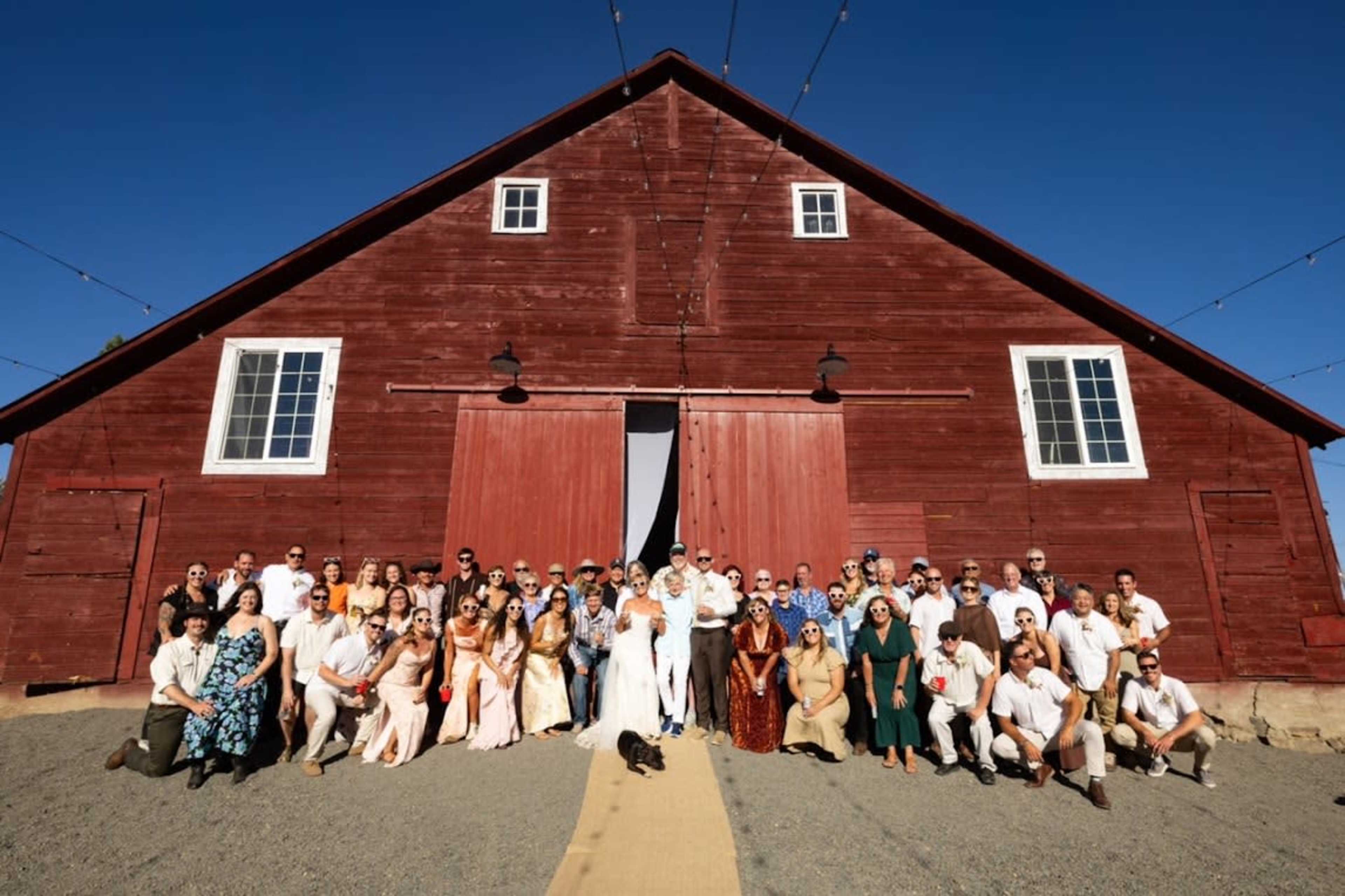A large group of people stands in front of a red barn with white accents on a clear day.