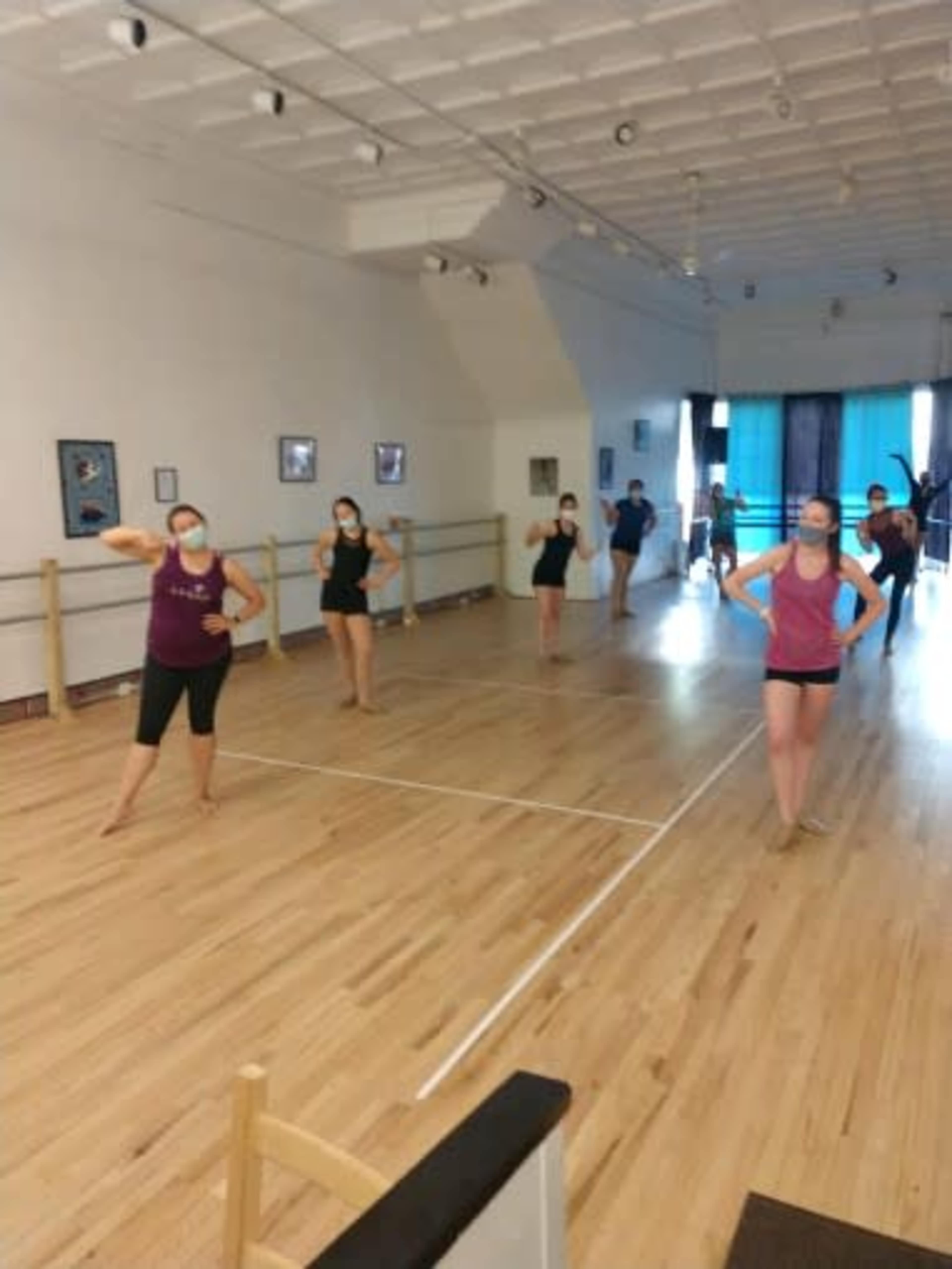A group of dancers in active pose rehearses in a wooden-floored studio with mirrors and a ballet barre.