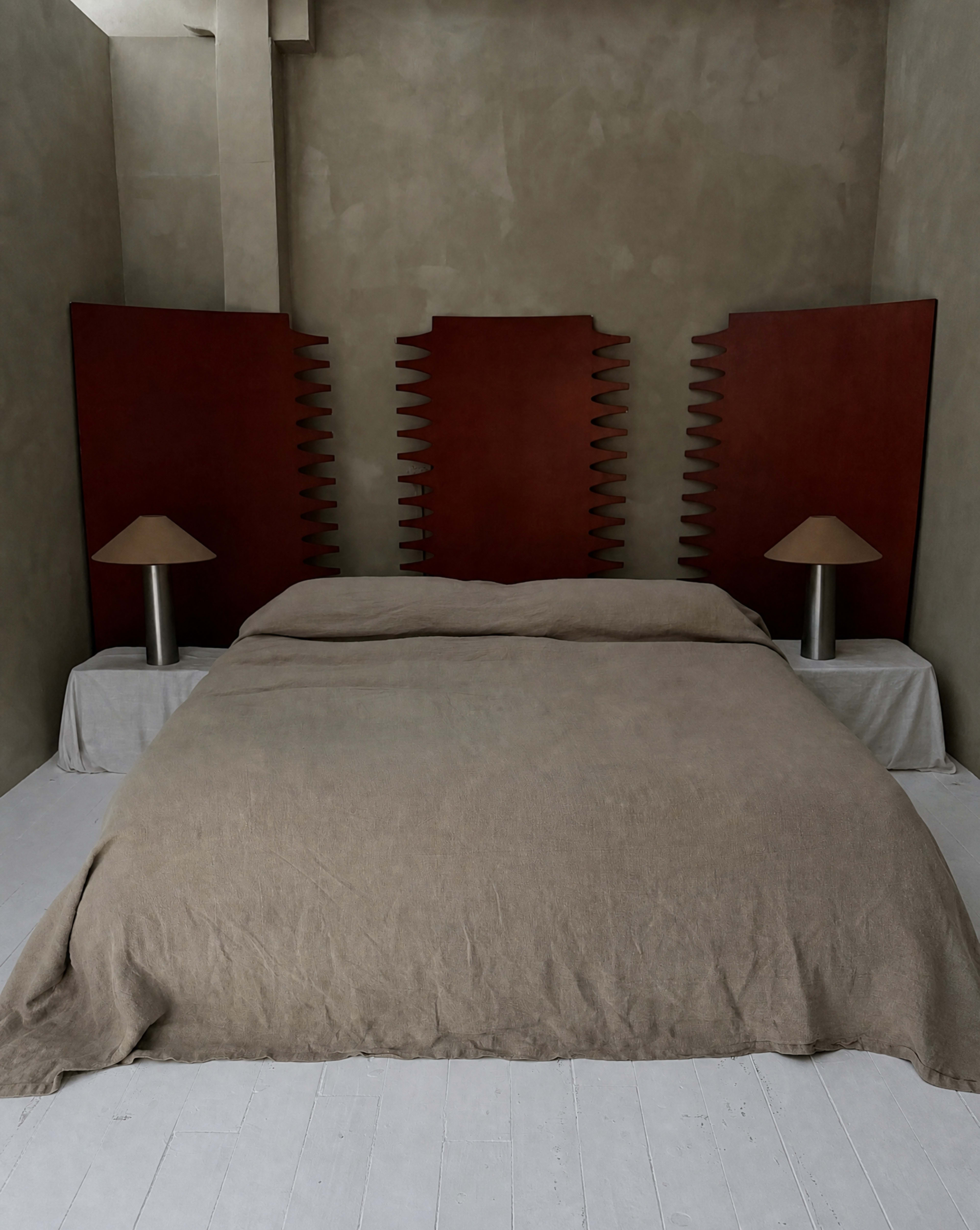 The image shows a minimalist bedroom featuring a large bed with a beige coverlet, flanked by two red wooden panels and lit by two simple lamps.