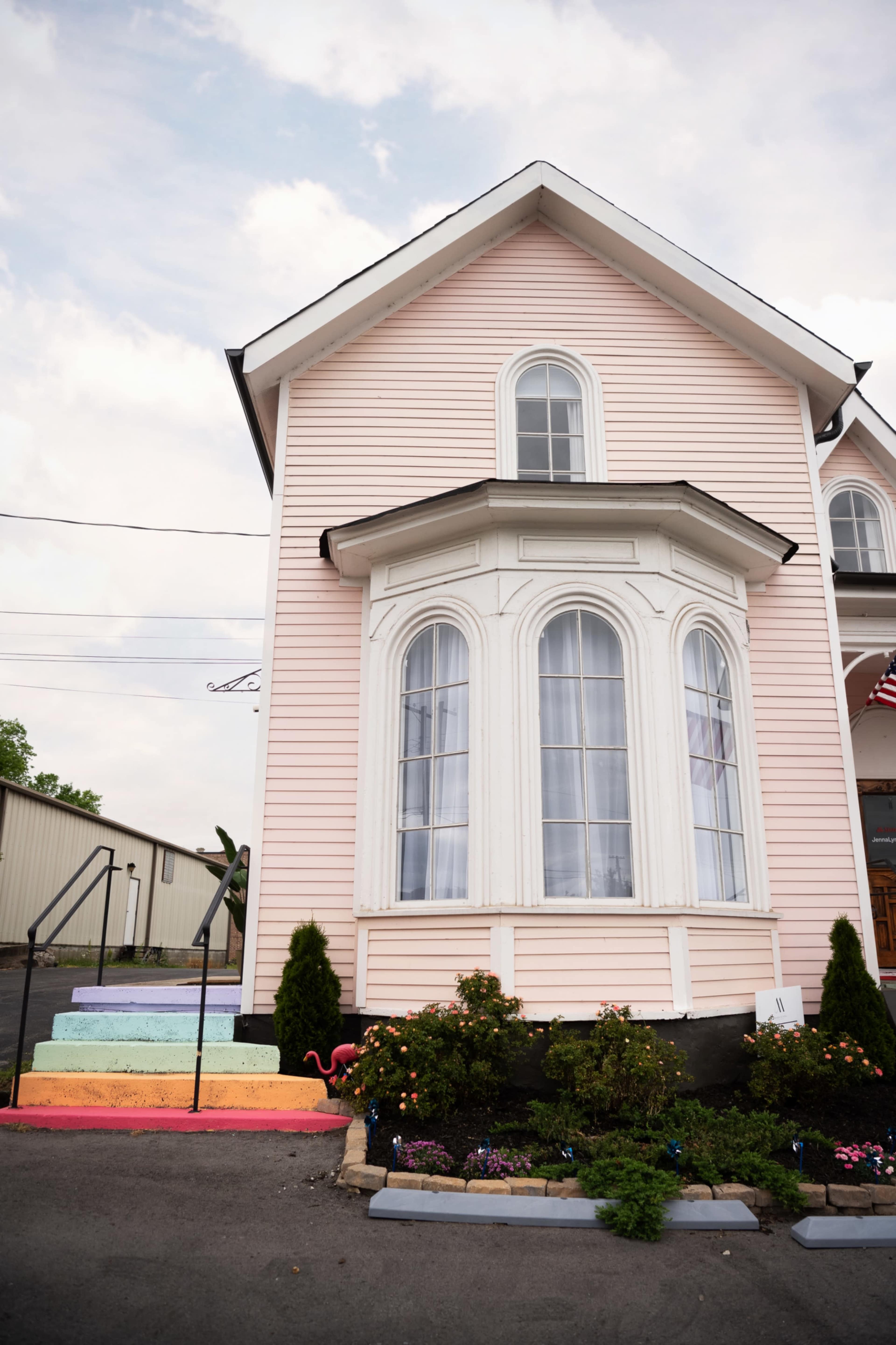 The image shows a pink house with a large bay window and colorful steps leading to a garden area with flowers.