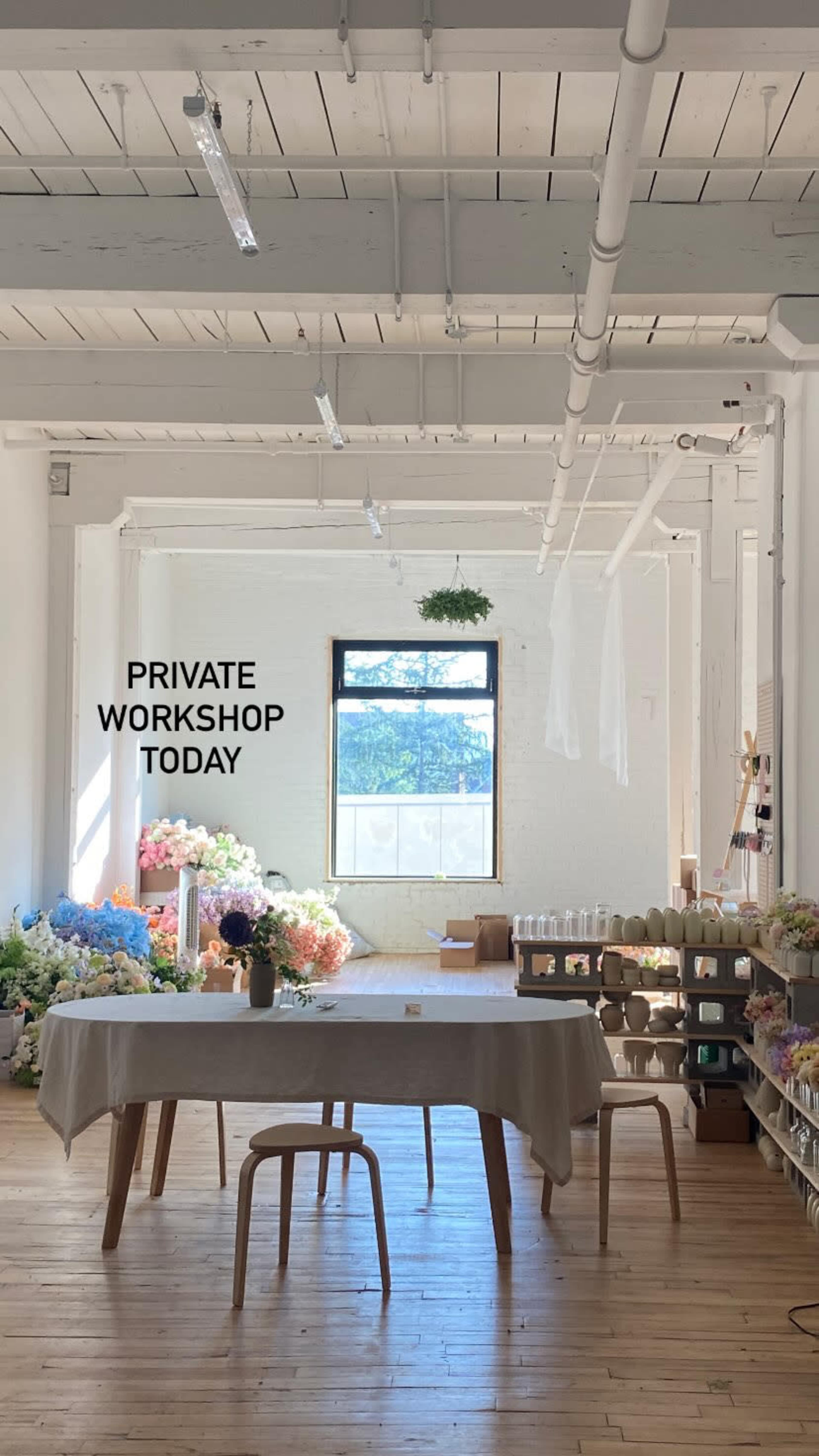 The image shows a bright, airy workshop space with a wooden table and stools, surrounded by floral arrangements and decor.