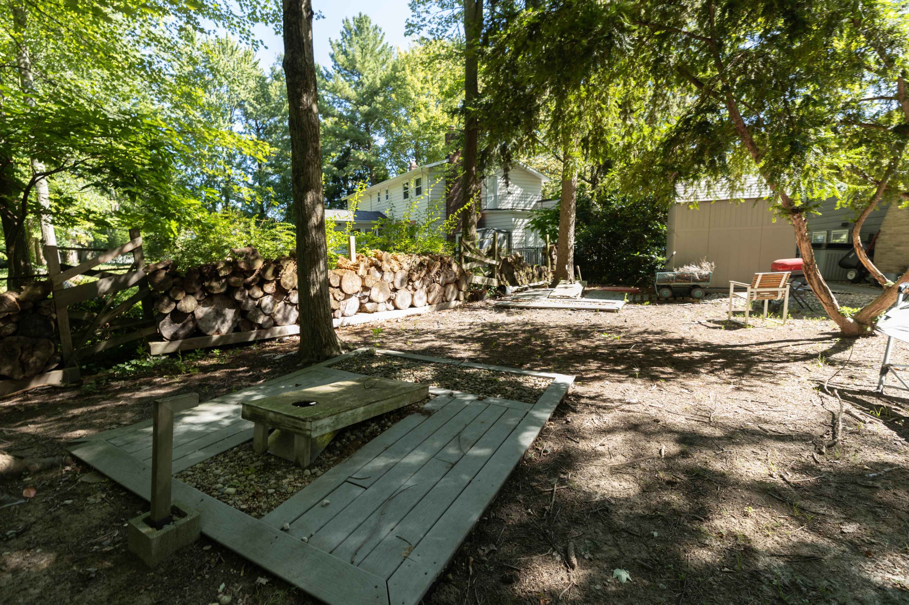 The image shows a yard with wooden platforms, a log pile, and a nearby house surrounded by trees.