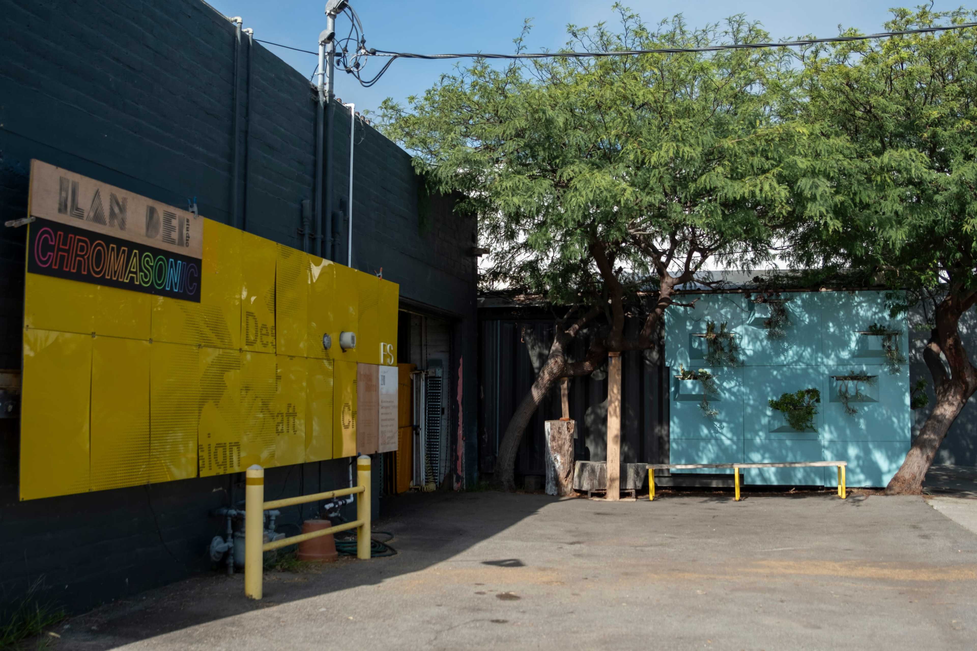 The image shows an outdoor space featuring two buildings with colorful facades, one yellow and the other blue, alongside a tree and utility poles in a paved area.