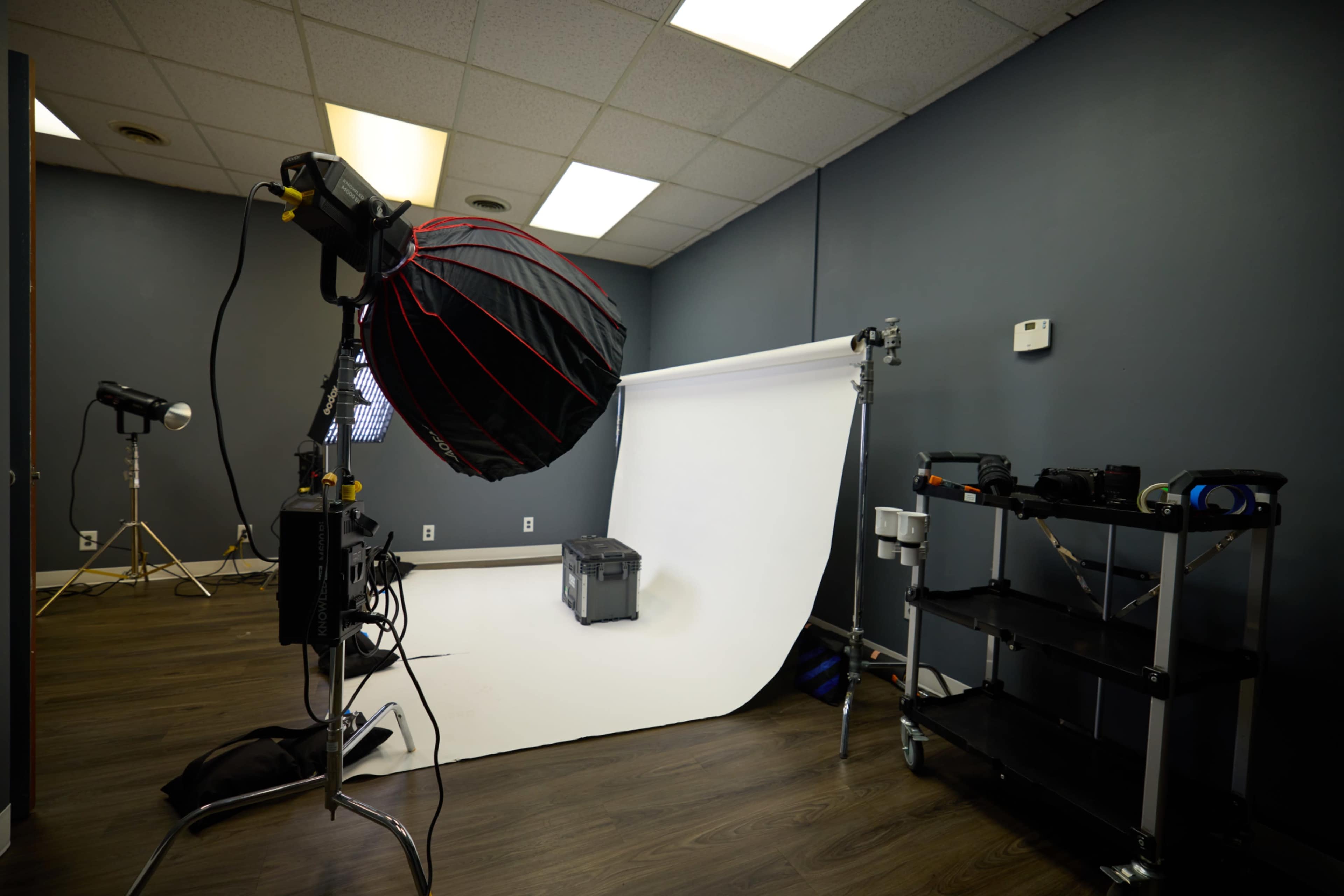 A photography studio features a white backdrop, softbox lighting, and various equipment on rolling carts.