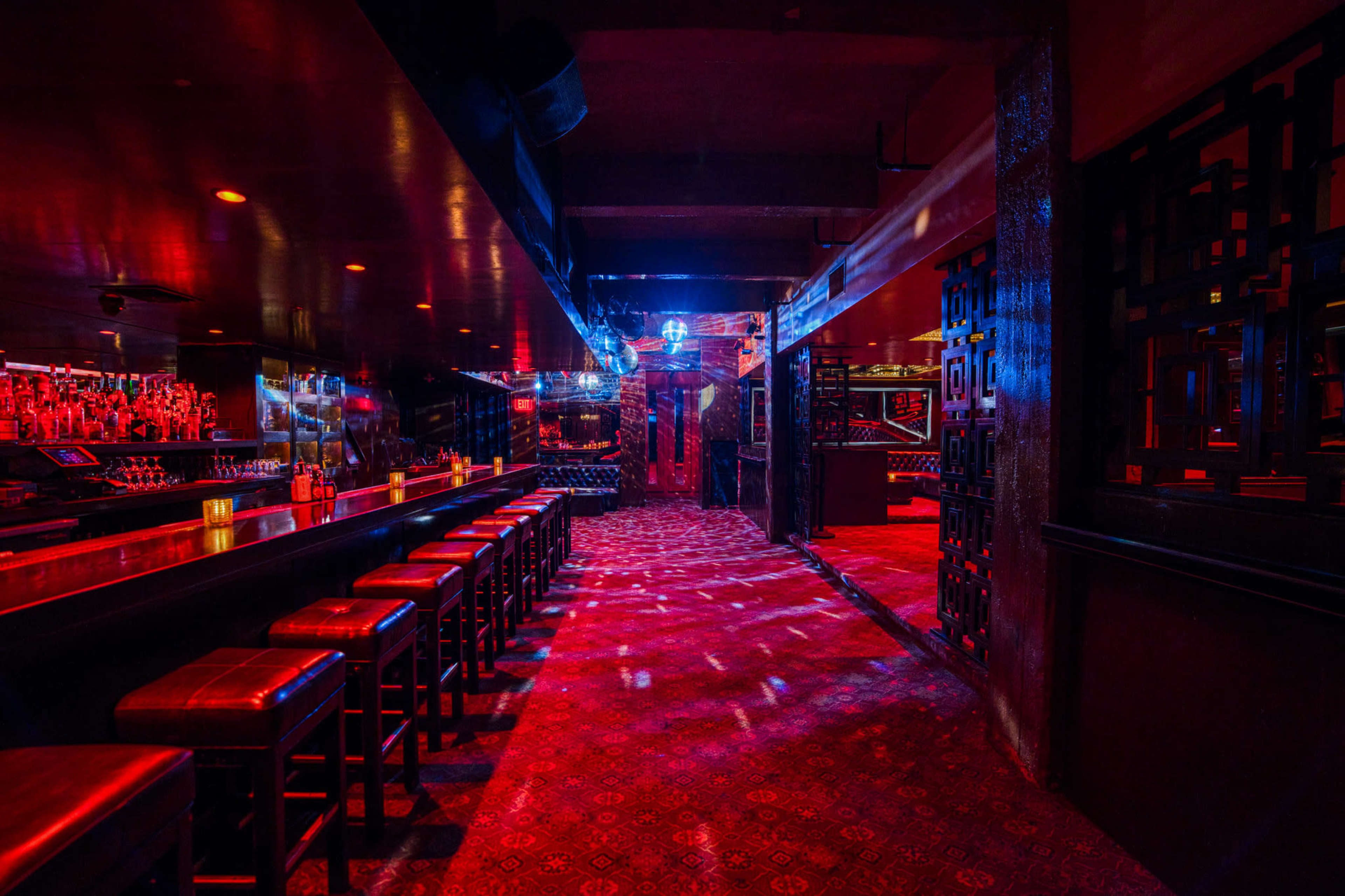 The dimly lit interior of a bar features a long counter with high stools and patterned carpeting, illuminated by colorful lights.