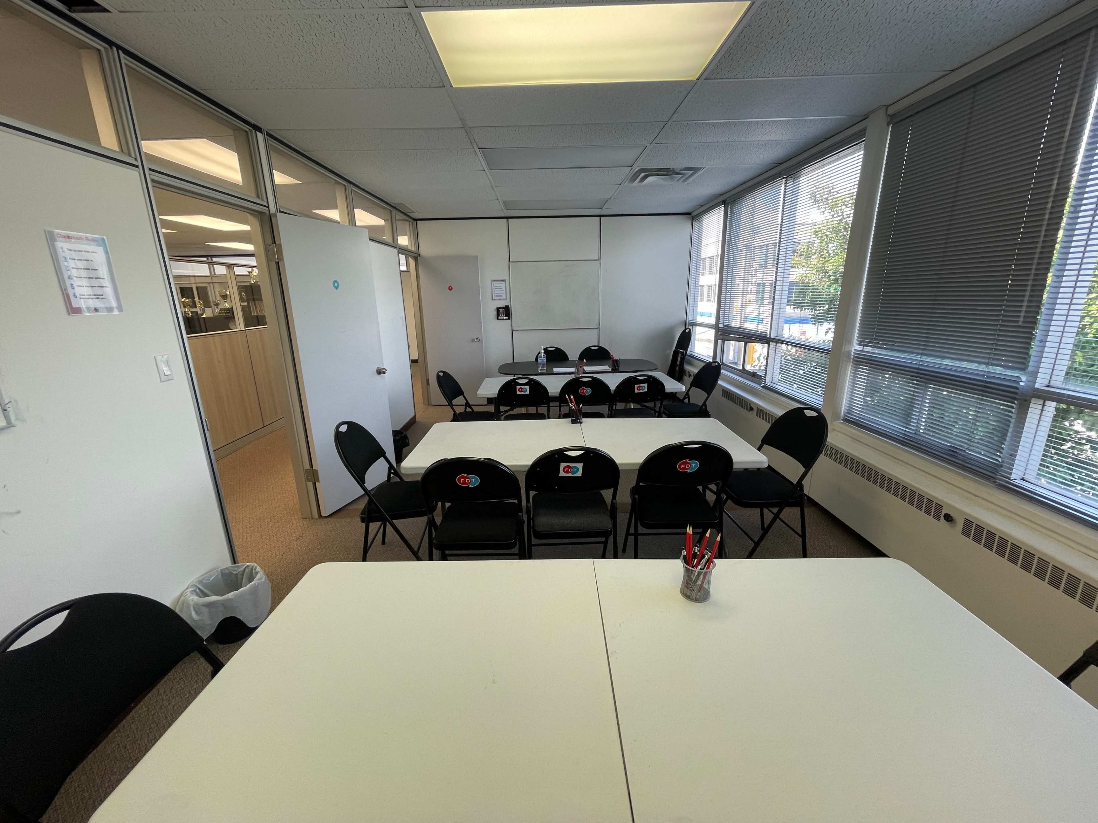 A classroom setting with four tables arranged in a rectangular formation and several black chairs placed around them, along with a few pen holders on the tables.