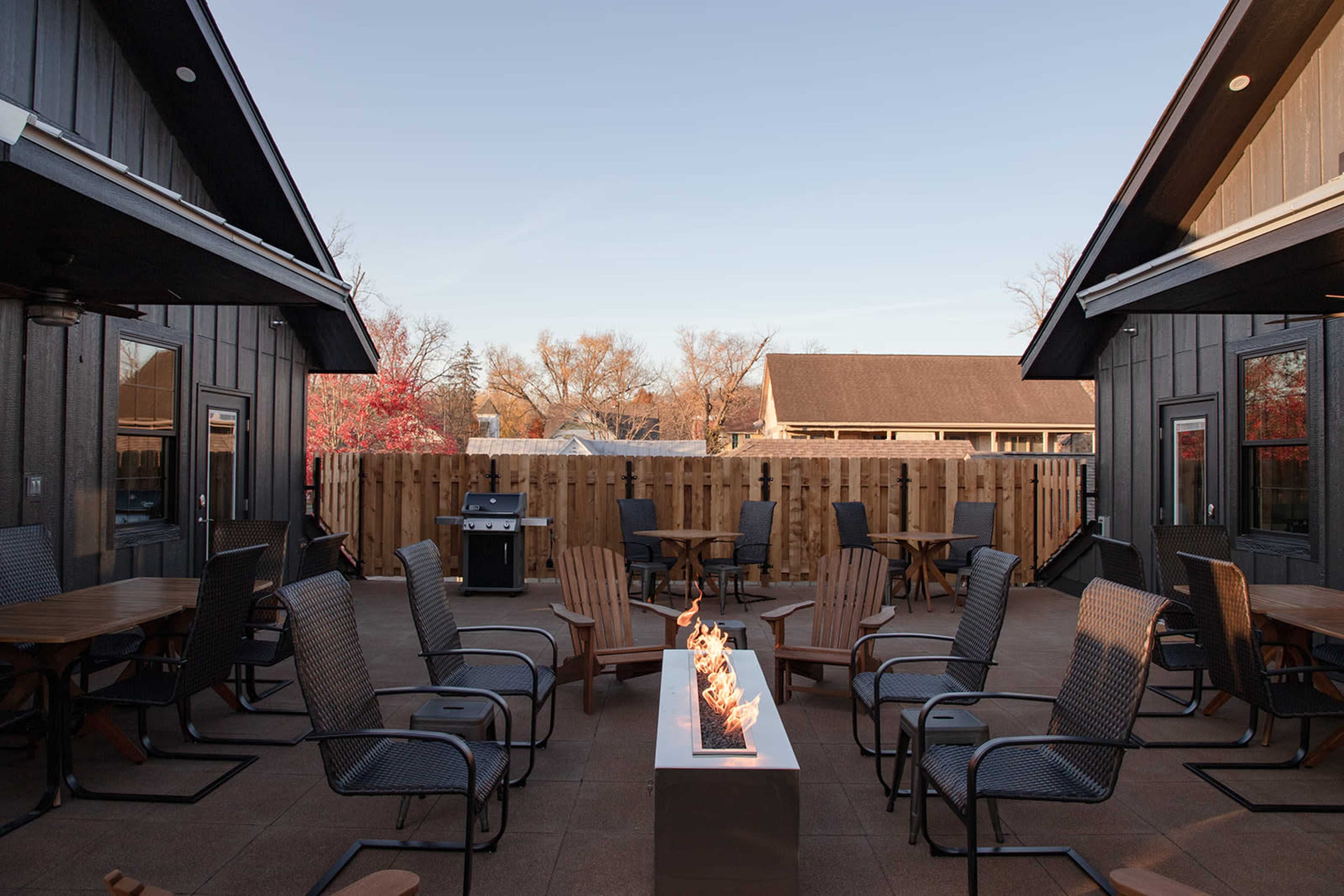 A patio area features several chairs arranged around a fire pit, with a grill and wooden fencing in the background.