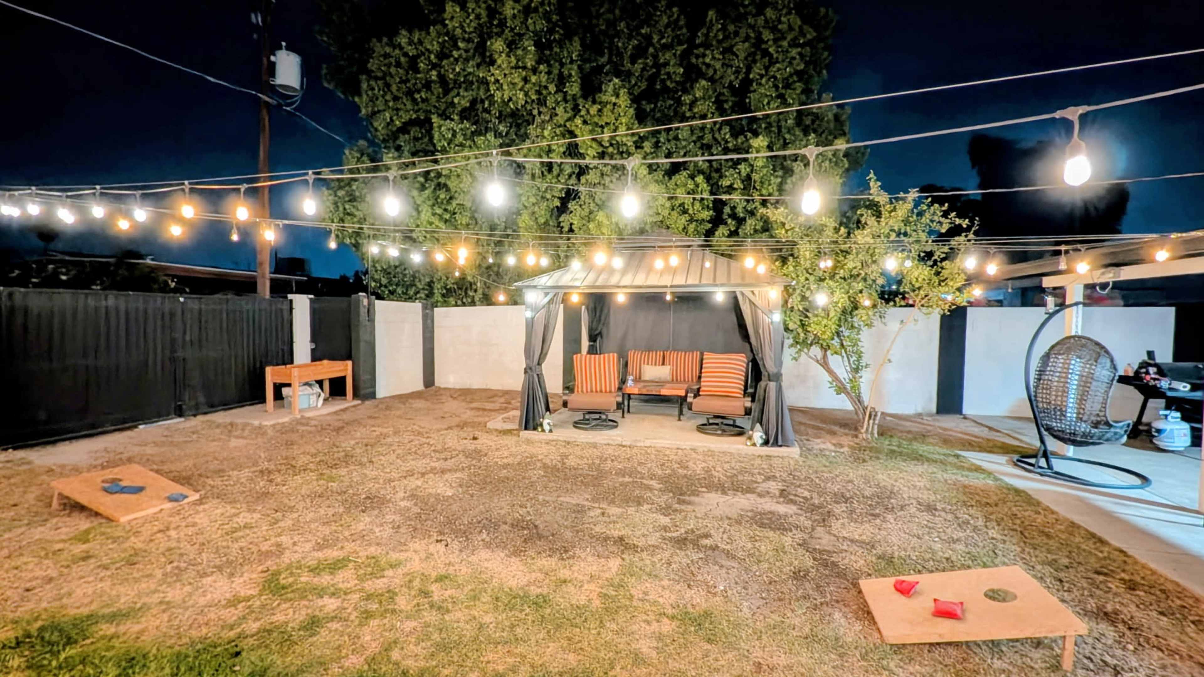 A backyard at night features a covered seating area with striped chairs, string lights overhead, and cornhole boards on the grass.