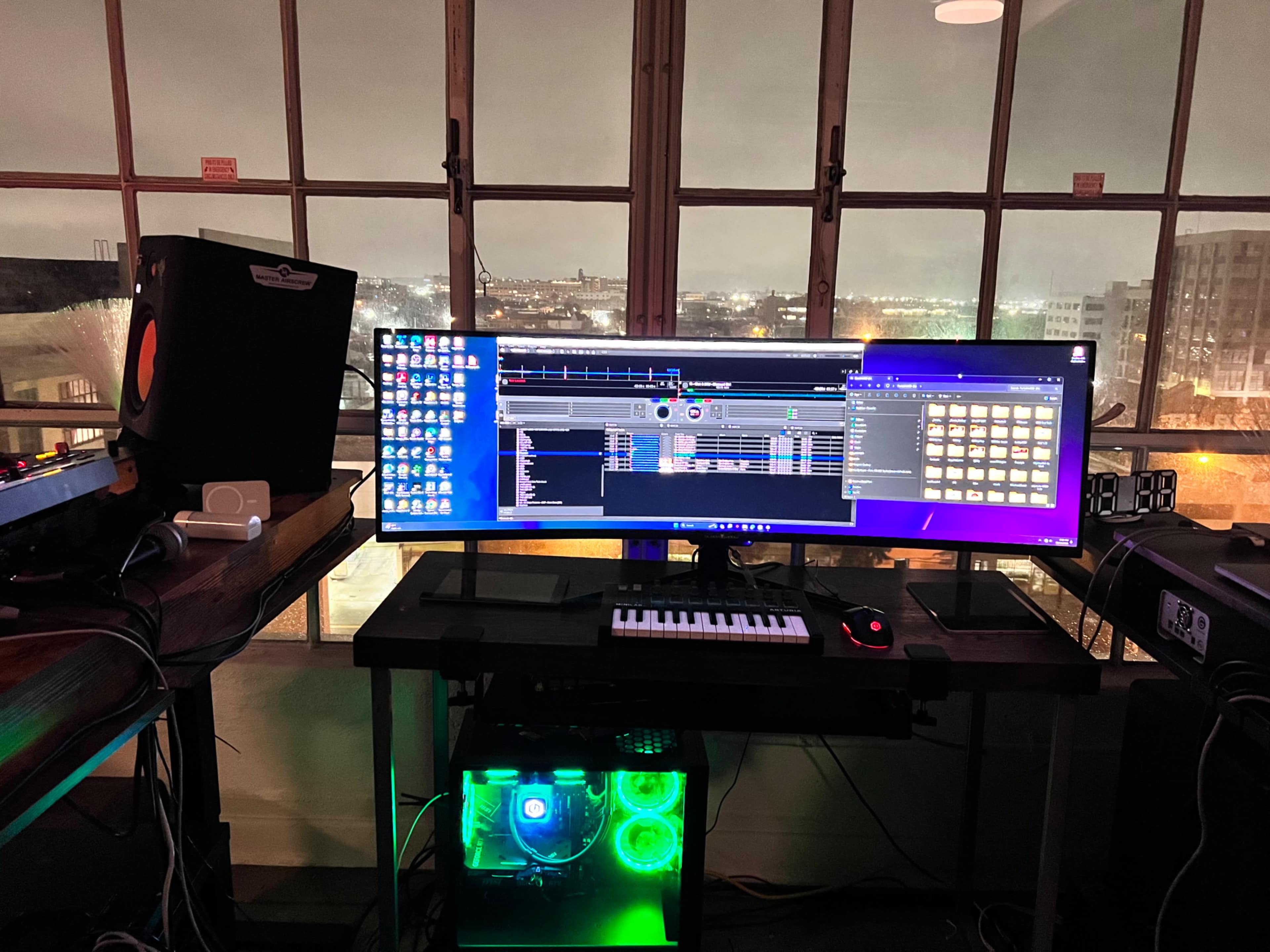 A computer workstation with dual monitors displaying digital audio software, surrounded by music production equipment and a city skyline visible through large, lit windows.