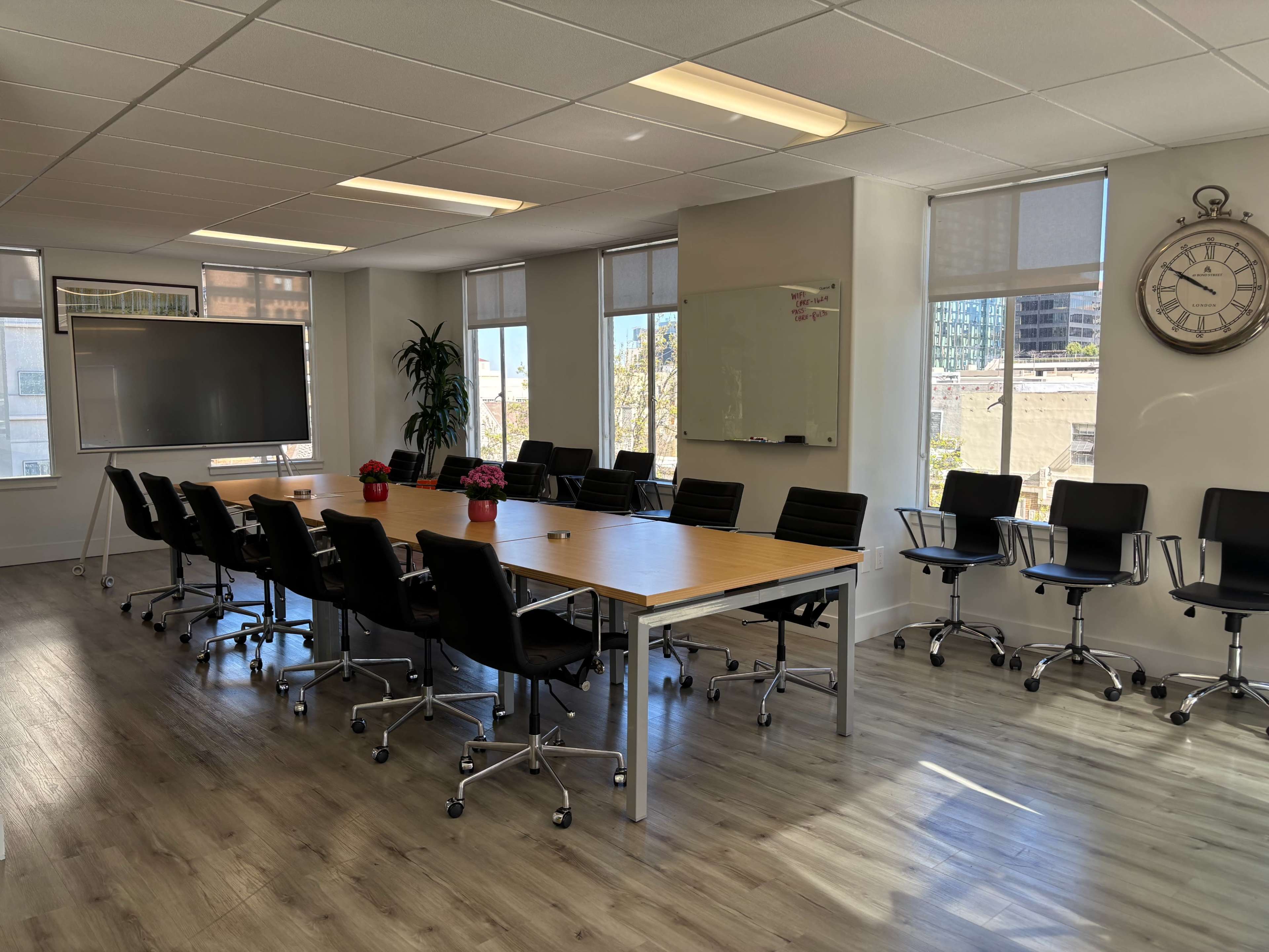 The image shows a modern conference room with a long wooden table surrounded by black chairs, large windows, a whiteboard, and a clock on the wall.