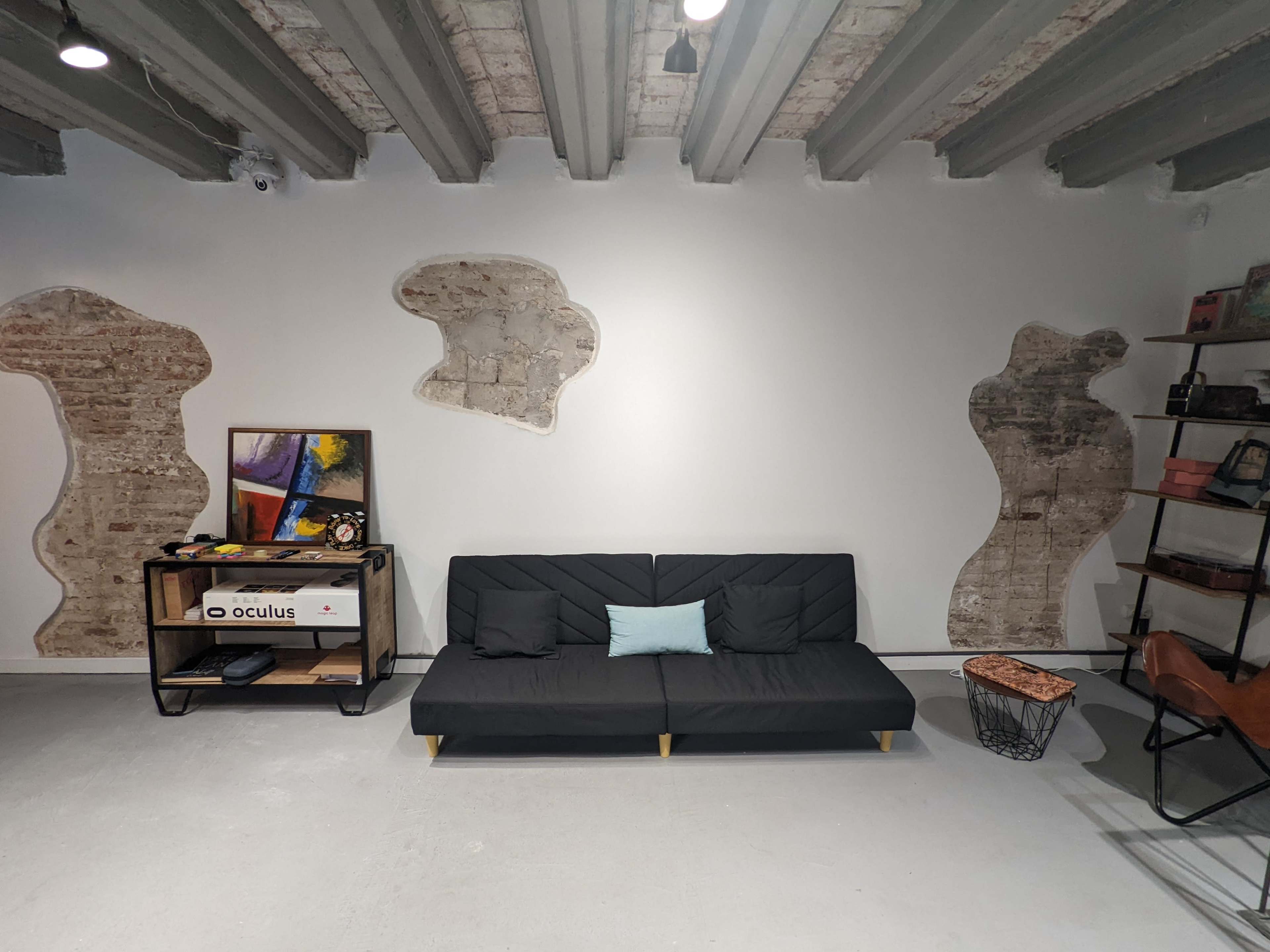 The image shows a modern living space featuring a black sofa with a light blue cushion against a wall with abstract-shaped plaster remnants and a metal shelf beside it.