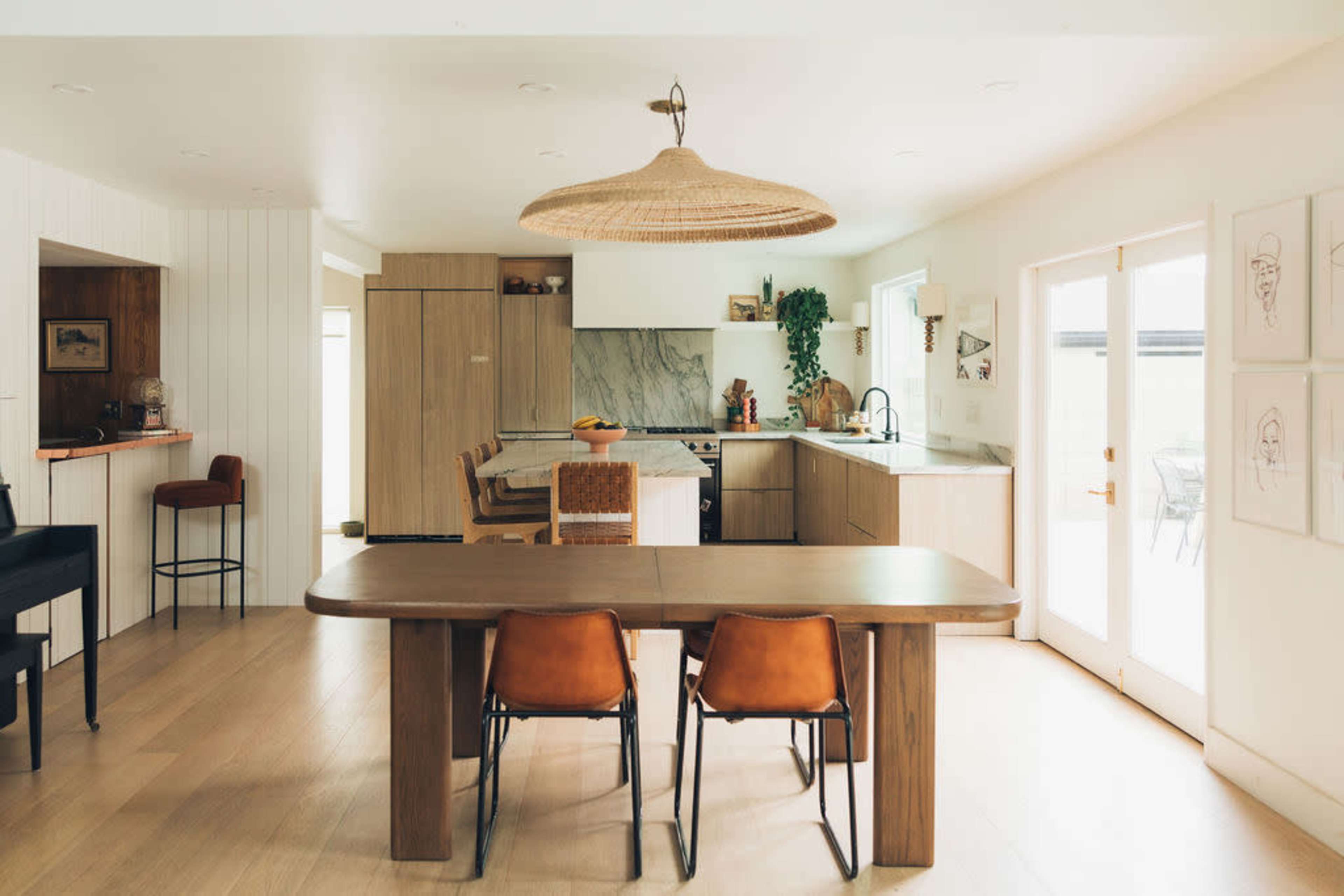 A modern kitchen and dining area featuring a wooden table, stylish chairs, and a large pendant light, with a mix of cabinetry and a marble backsplash in the background.