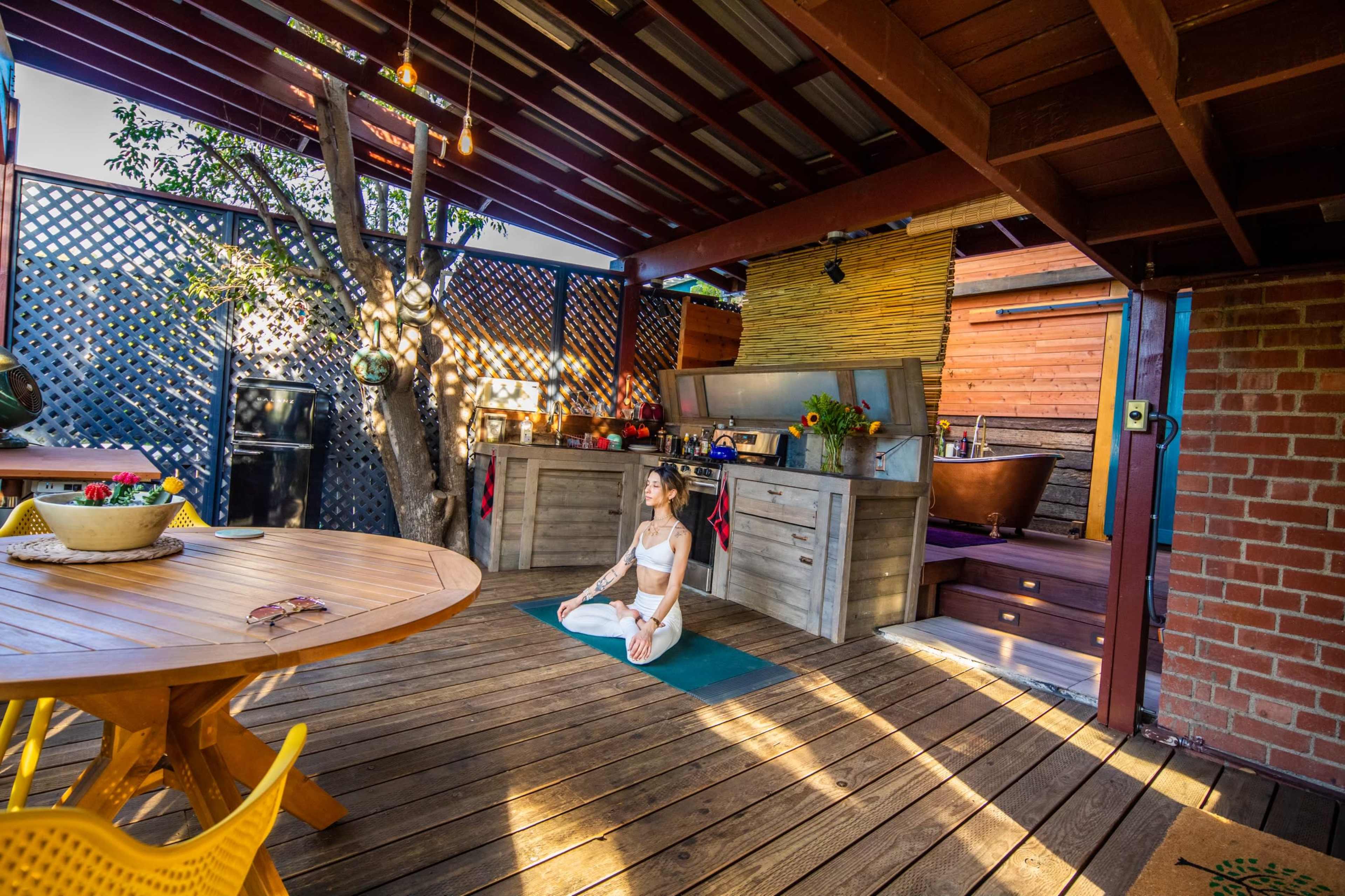 A woman practices yoga on a mat in a wooden deck area with an open kitchen and plants surrounding her.
