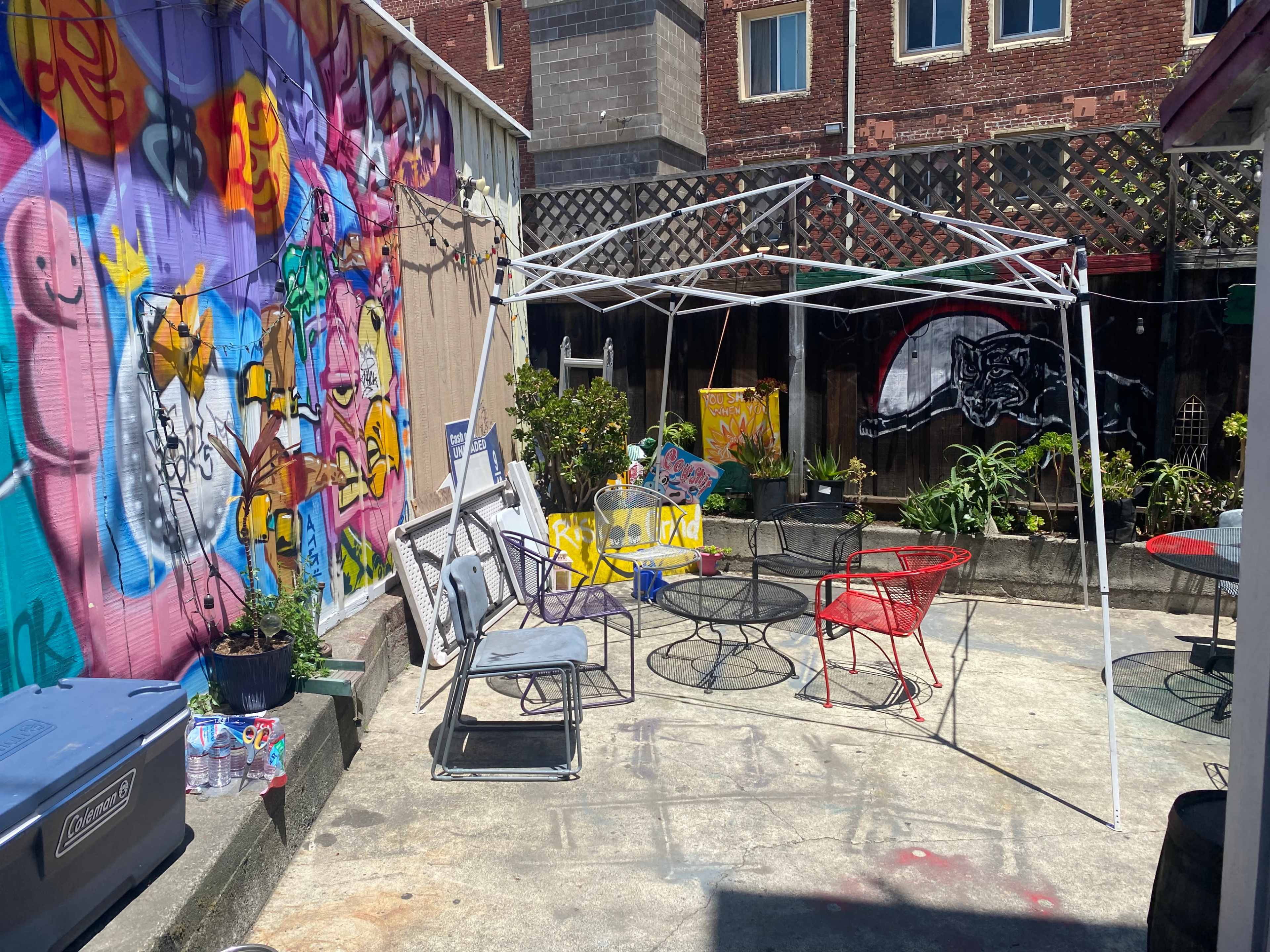 The image shows an outdoor space with a white canopy, several chairs, and colorful murals on the walls.