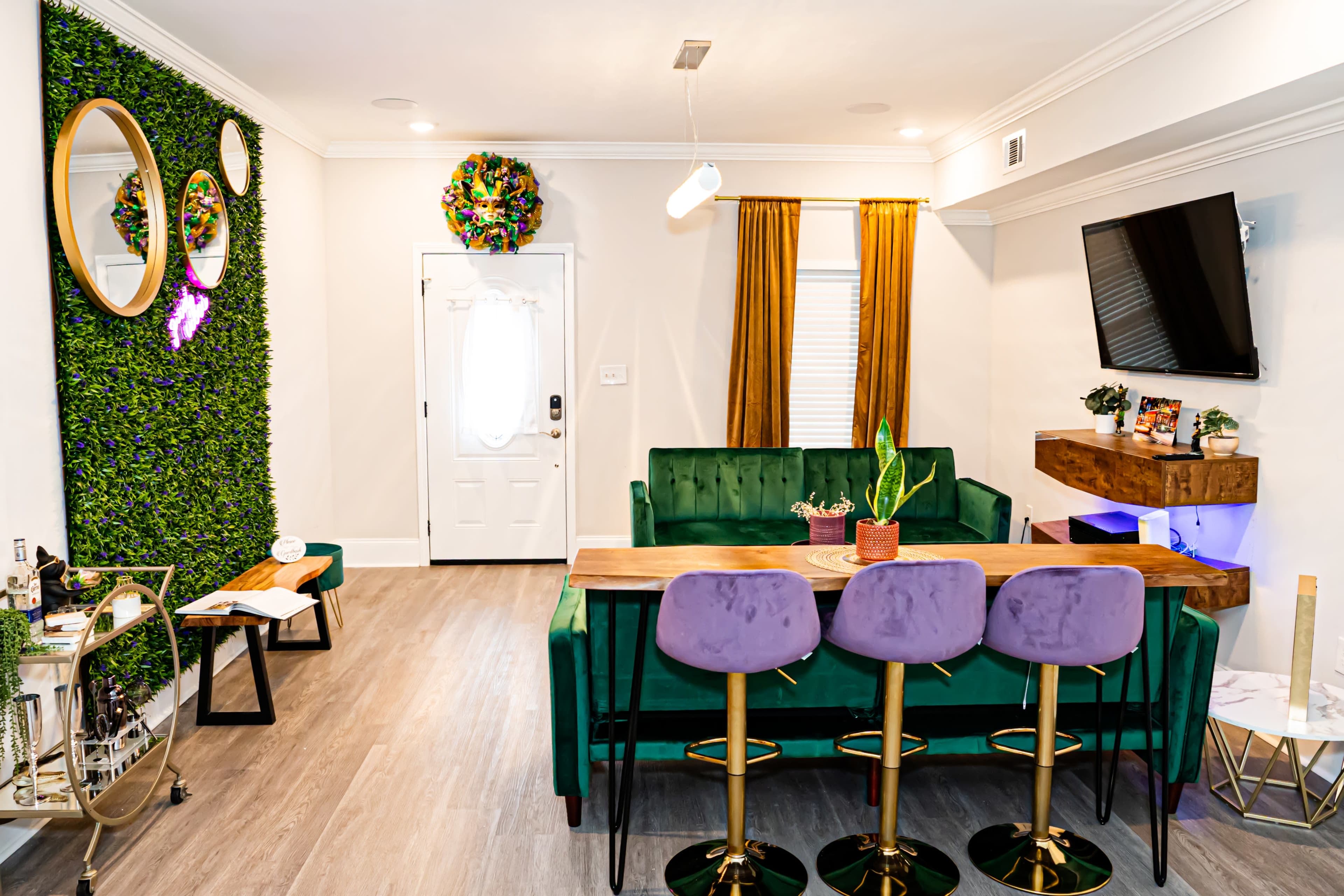The image shows a modern living space with a green sofa, a wooden dining table surrounded by pink bar stools, a plant, and a decorative green wall with mirrors.