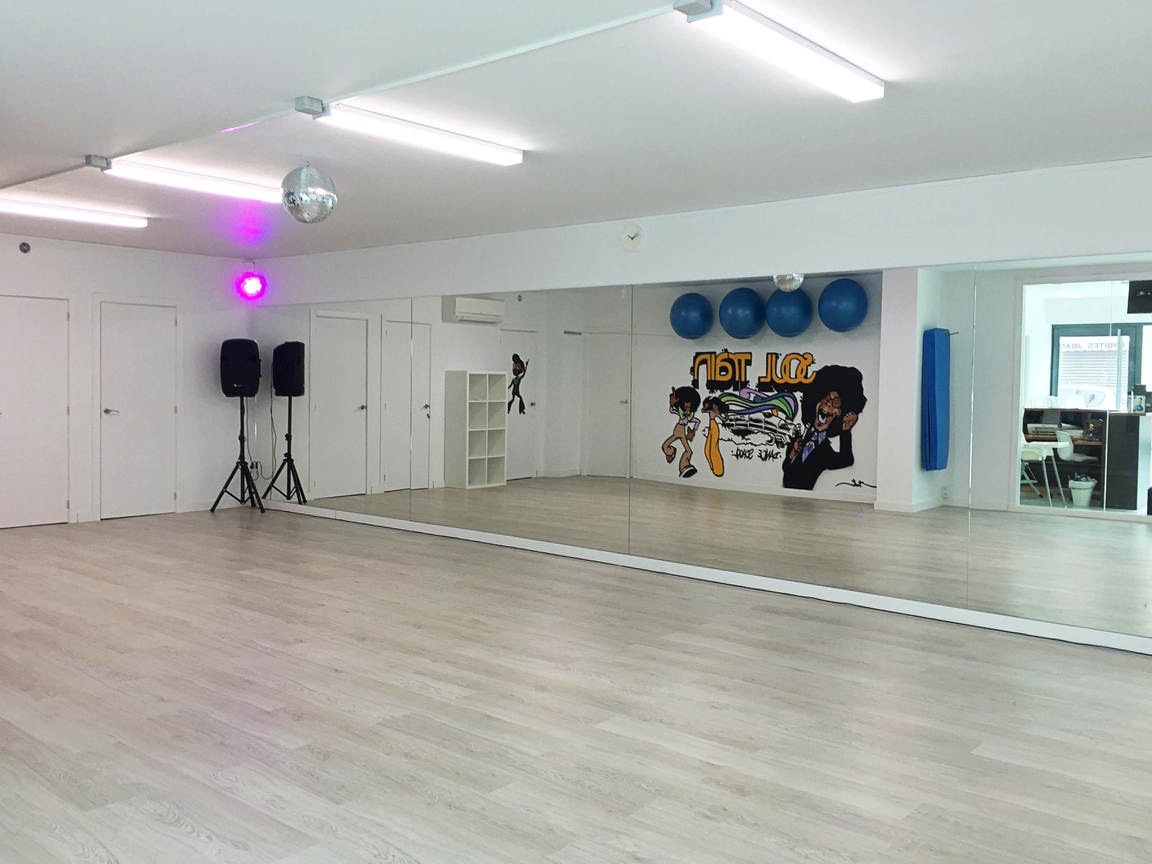 A dance studio with a mirrored wall, wooden flooring, and a colorful mural alongside blue exercise balls.
