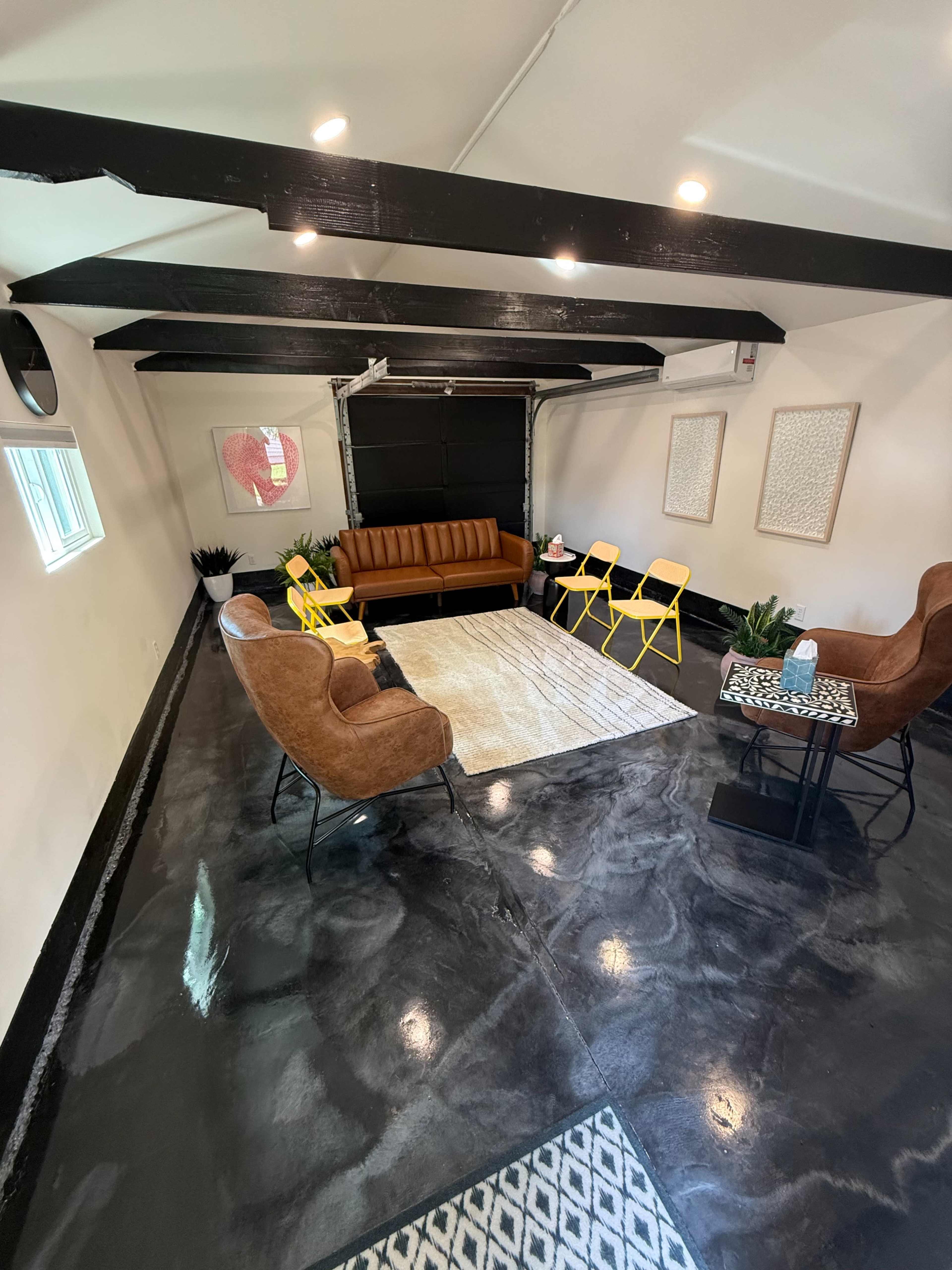 The image shows a modern indoor space with a mixed seating arrangement consisting of a brown couch, two armchairs, and yellow folding chairs, along with plants and artwork on the walls.