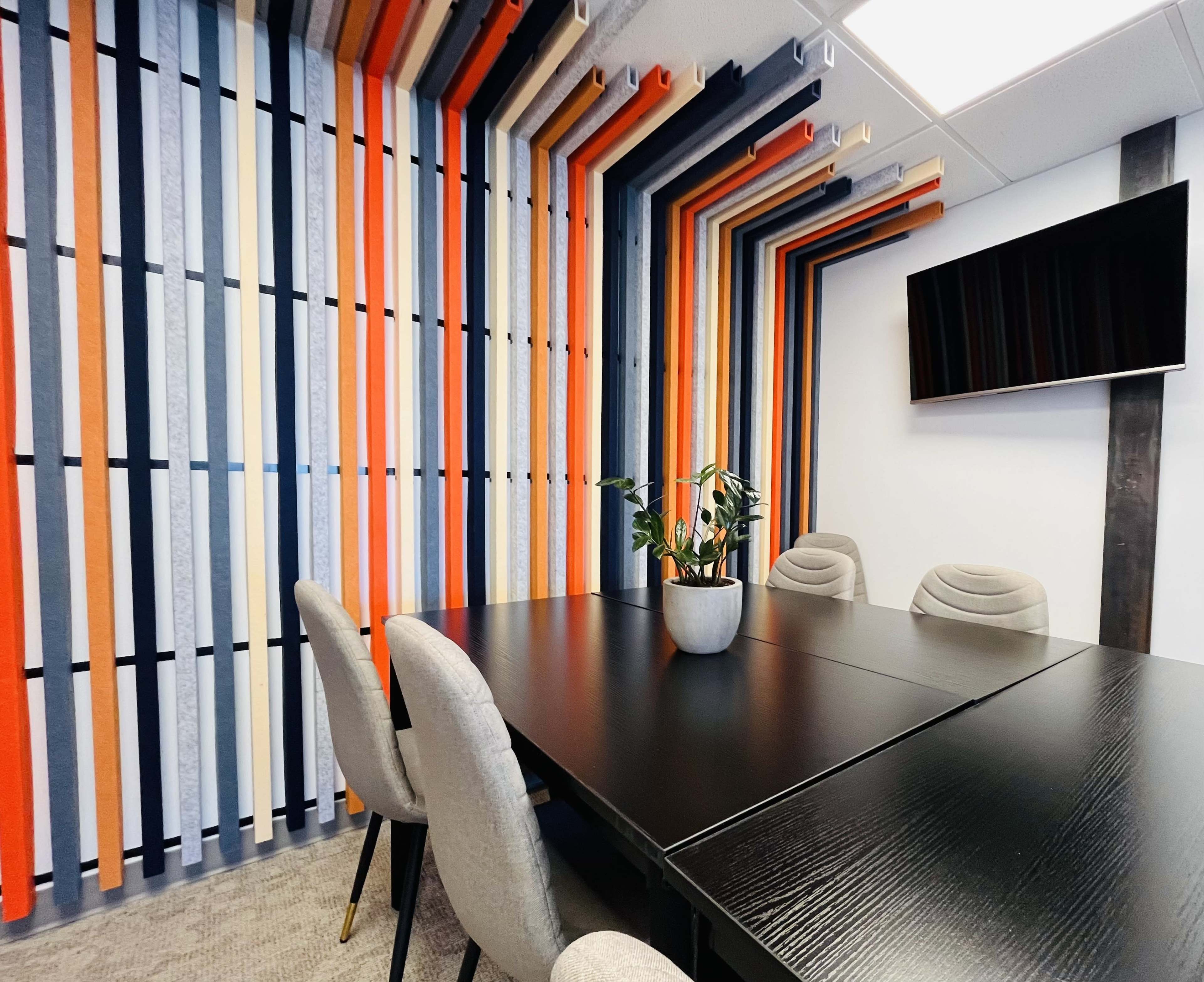 A conference room features a black table surrounded by gray chairs and a colorful striped wall, with a TV mounted on the wall and a plant in the center.