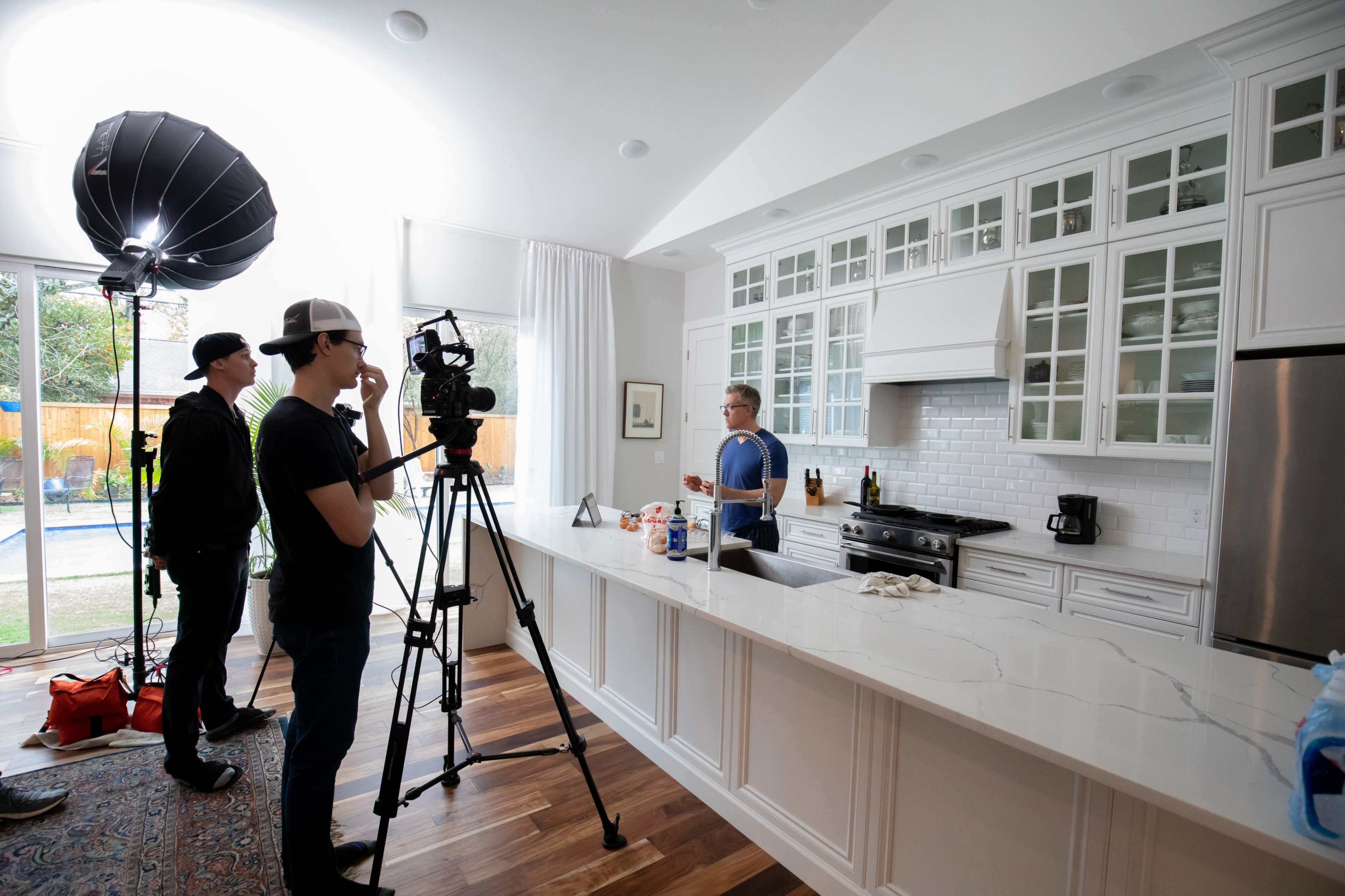 A film crew sets up to record a cooking segment in a modern kitchen with white cabinetry and a large island.