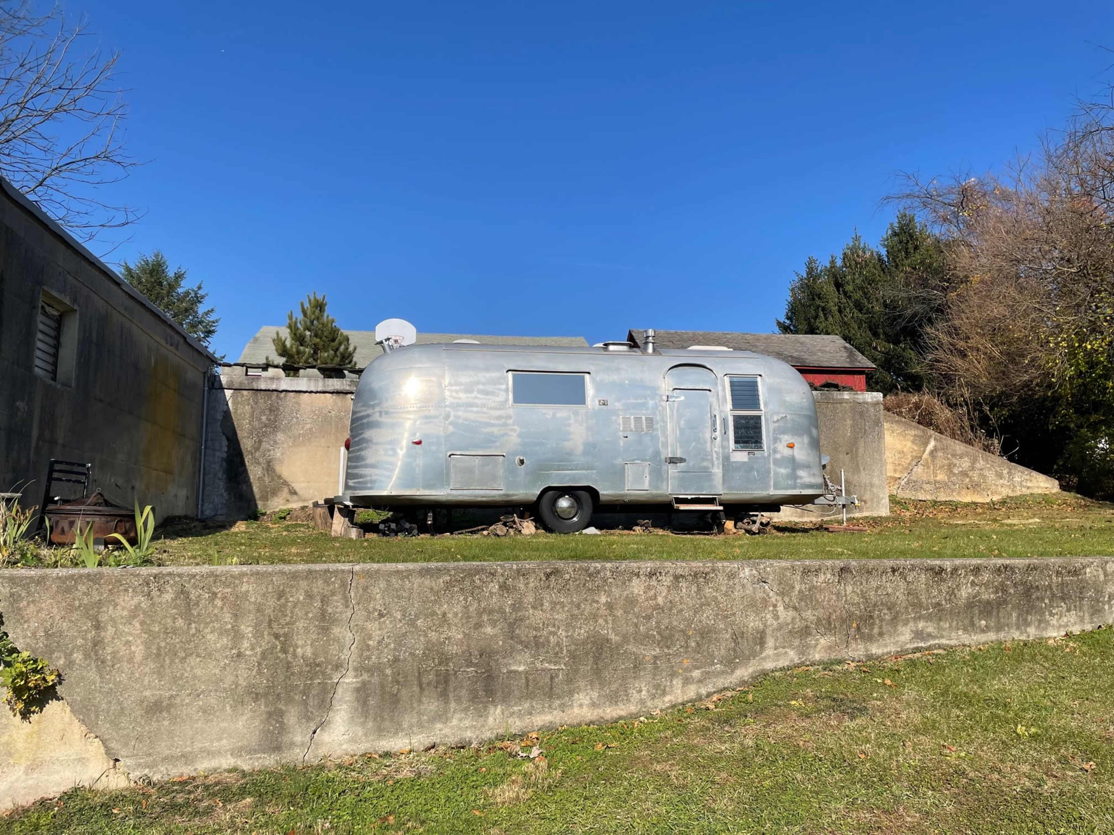 A vintage Airstream trailer is parked on a grassy area next to a concrete wall under a clear blue sky.