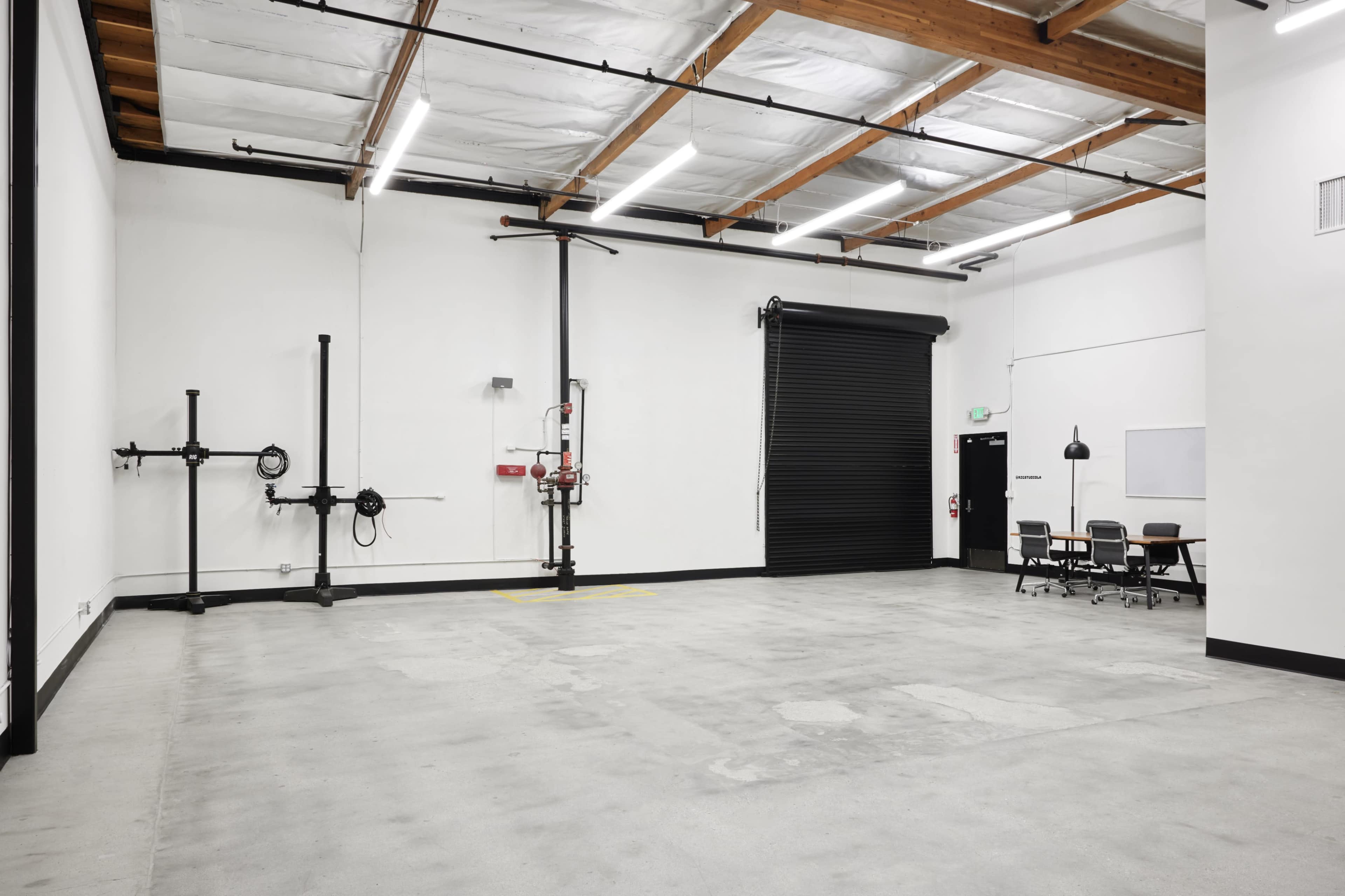 The image shows a spacious, empty industrial room with exposed wooden beams, a roll-up door, and a few pieces of simple furniture arranged against a wall.