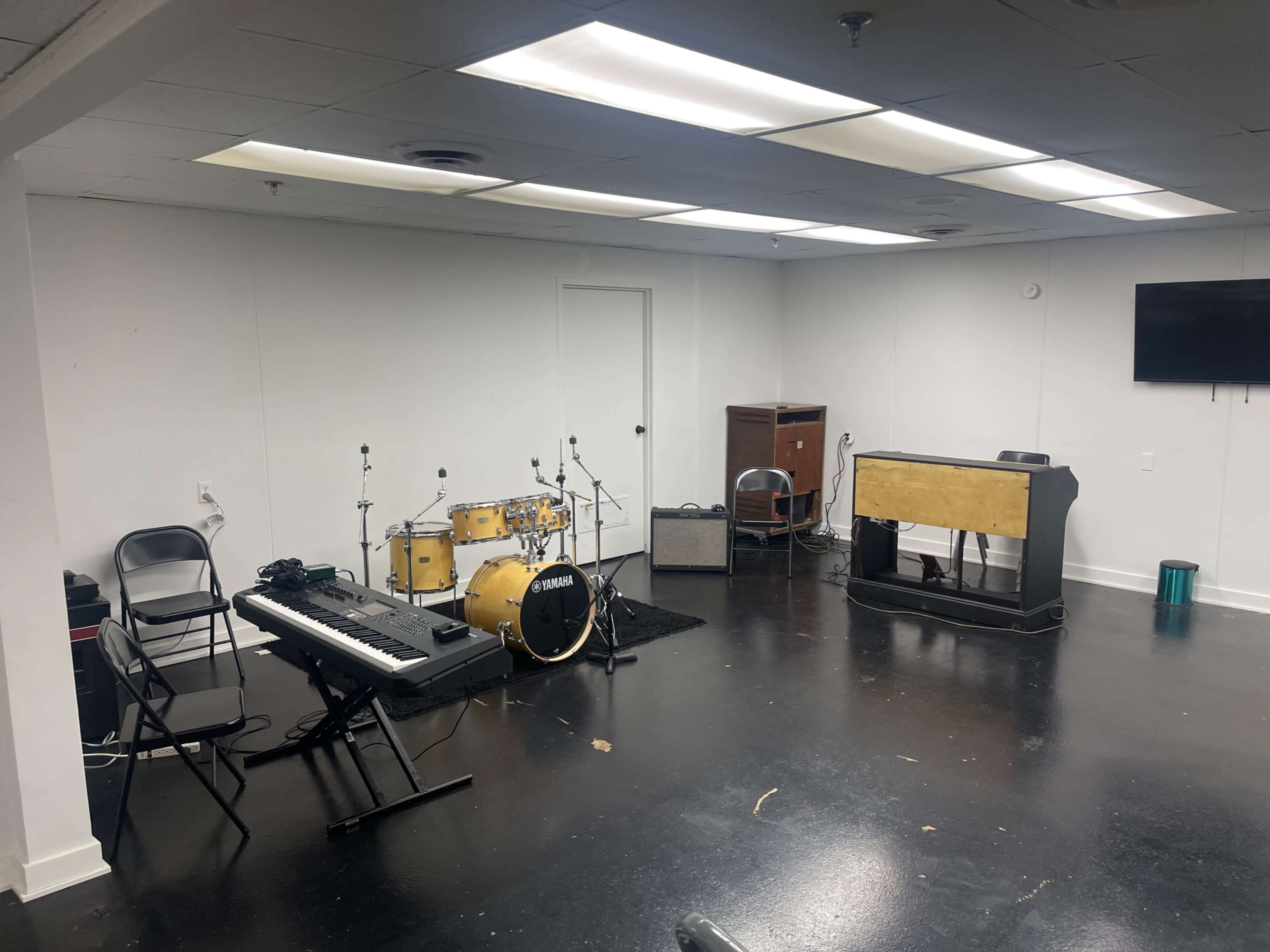 The image shows a blank room with a keyboard, a drum set, an amplifier, and an upright piano arranged around the space.