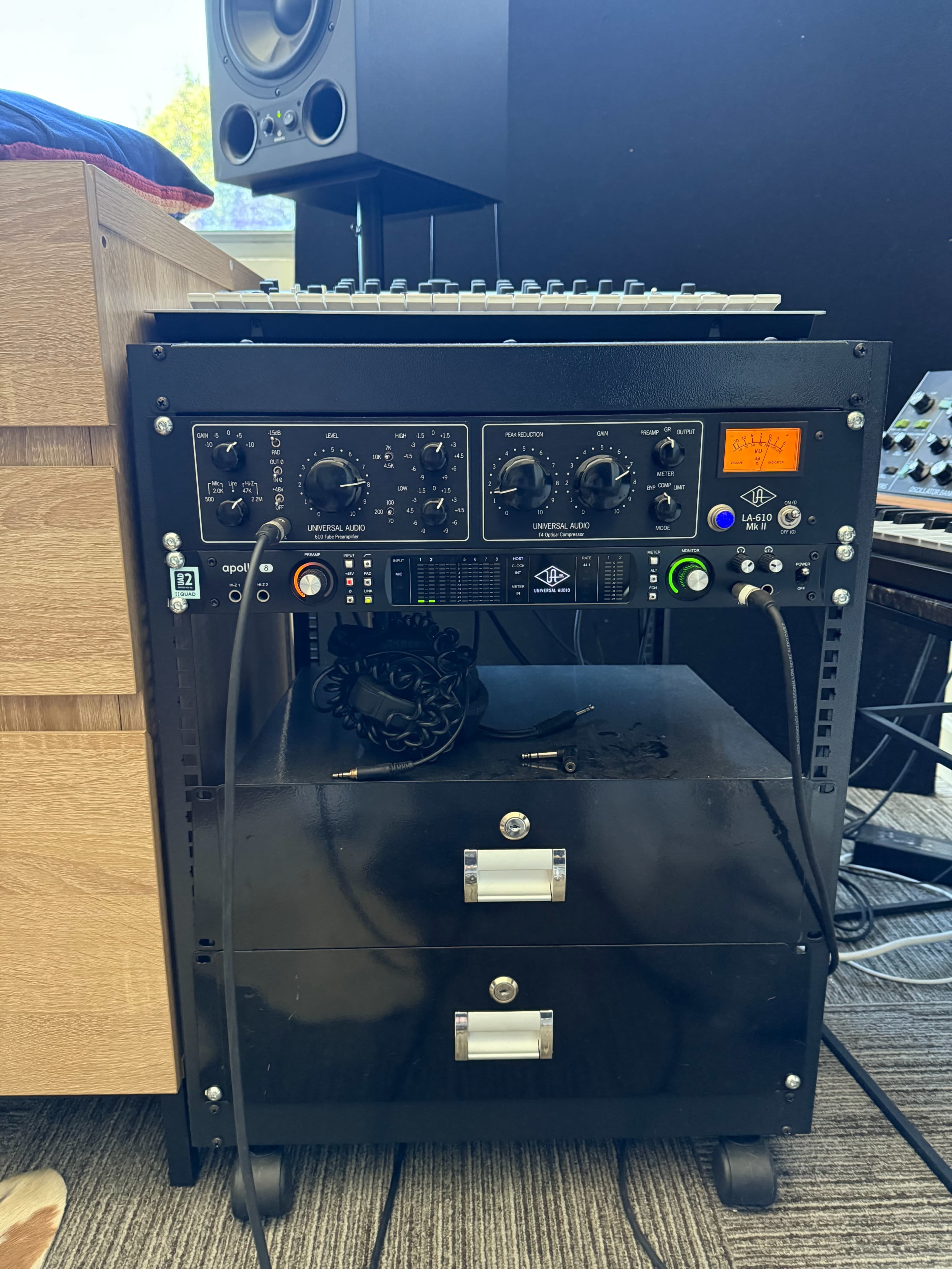 The image shows a piece of audio equipment with several knobs and meters, housed in a rack beneath a wooden surface, alongside a synthesizer and studio monitor.