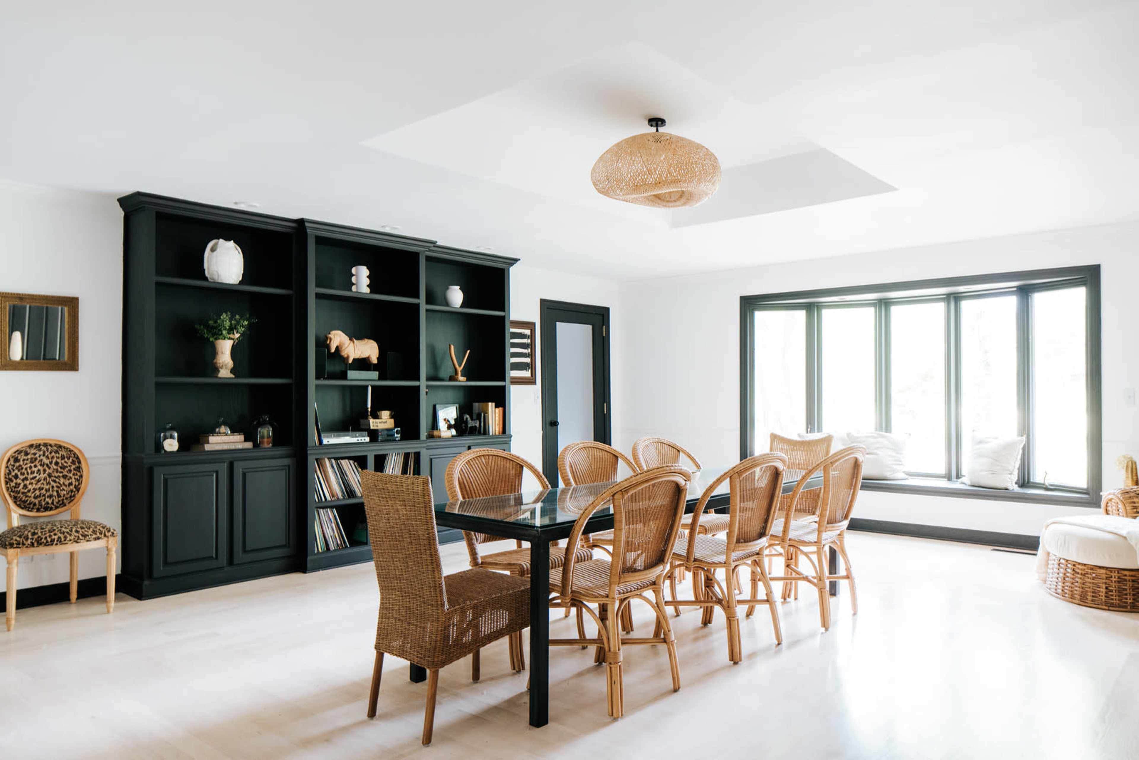 A dining area features a black bookcase, a glass table surrounded by woven chairs, and a large window seat with pillows.