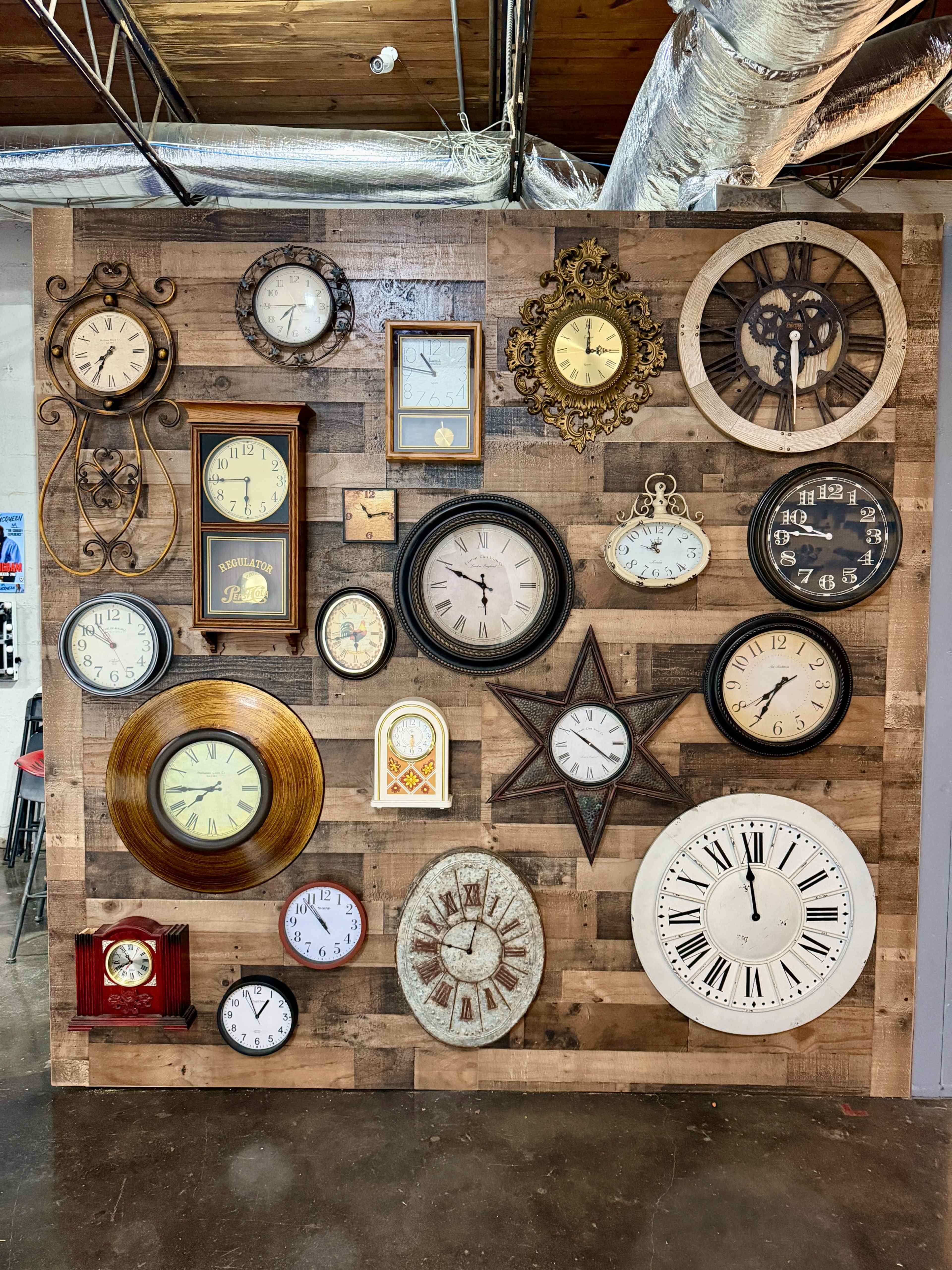 The image shows a collection of various clocks mounted on a wooden wall, featuring different styles and sizes.