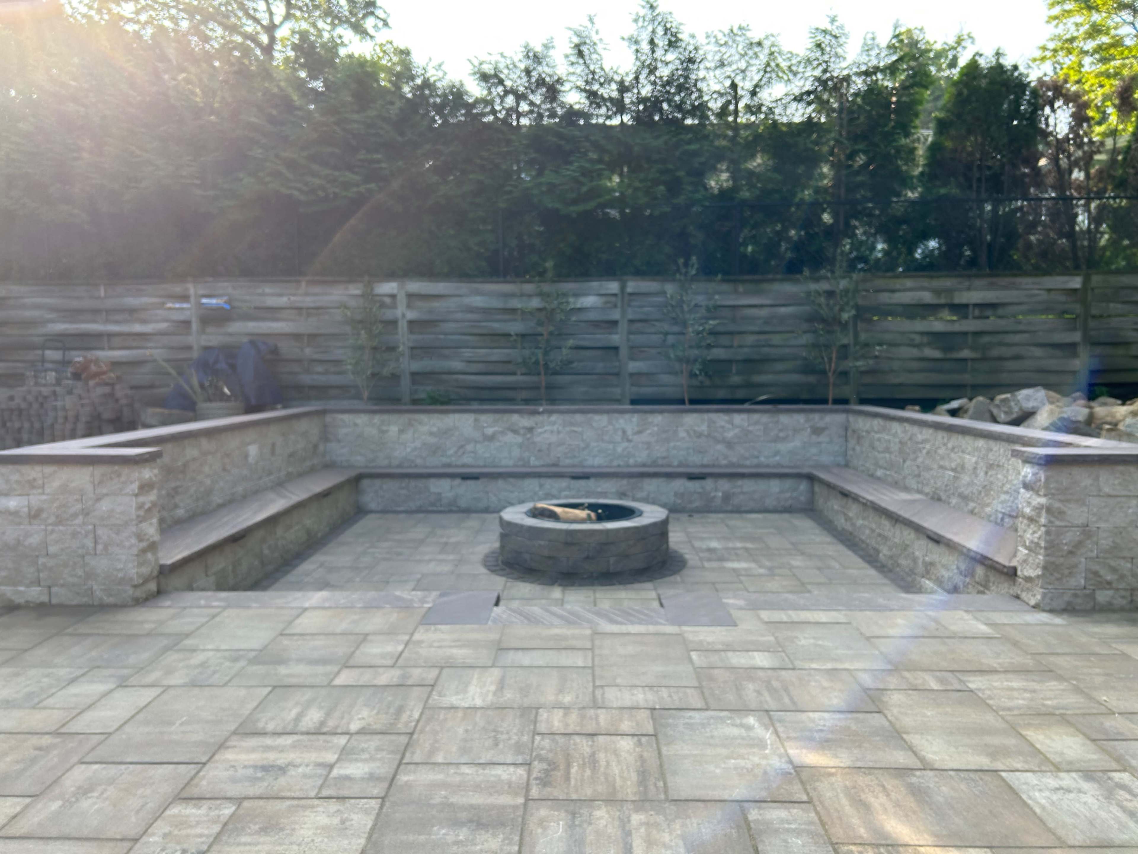 A stone patio with a central fire pit surrounded by built-in seating, enclosed by a wooden fence and greenery.
