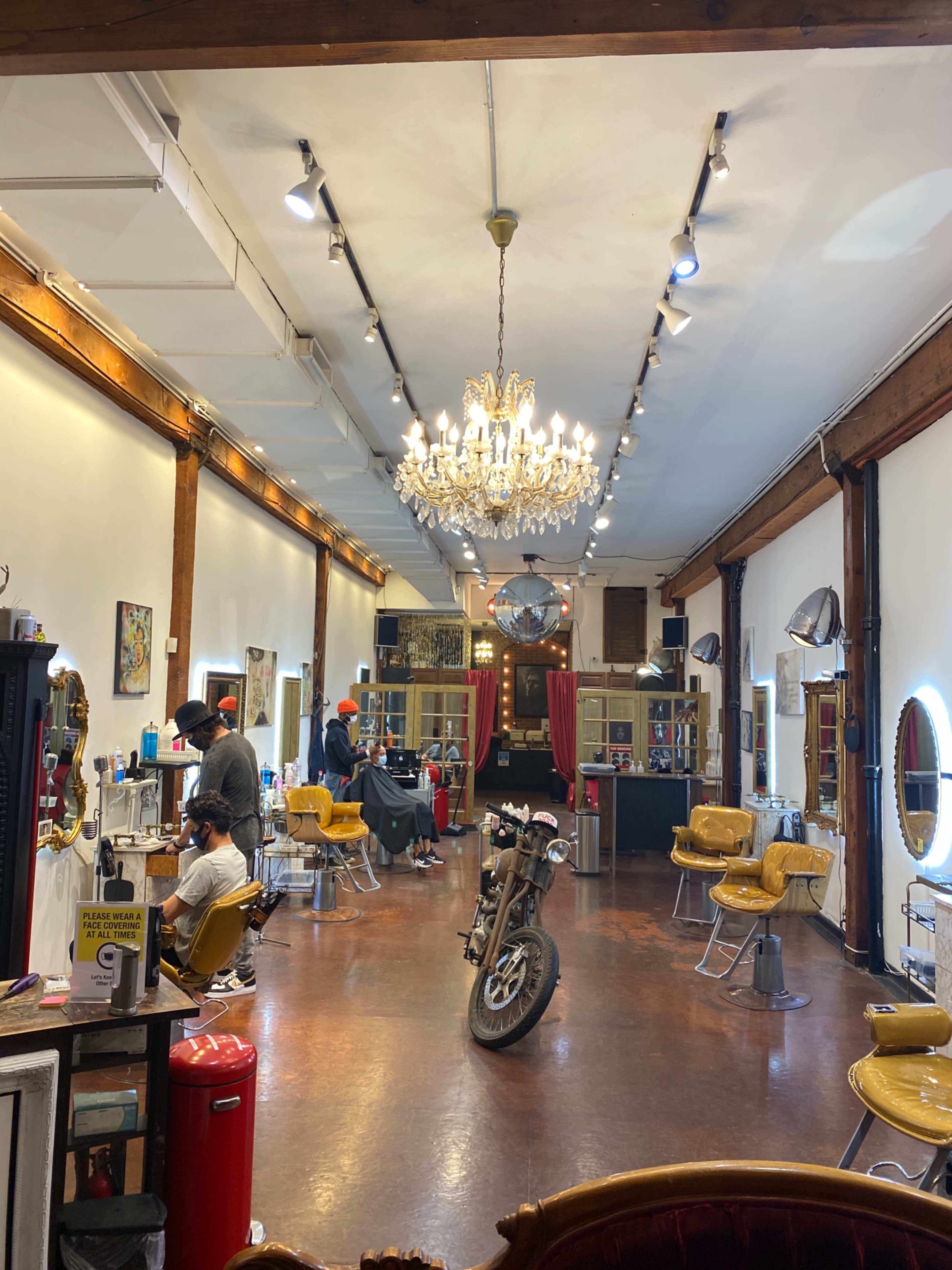 The interior of a barbershop features vintage yellow chairs, a chandelier, and a motorcycle positioned in the center of the floor.