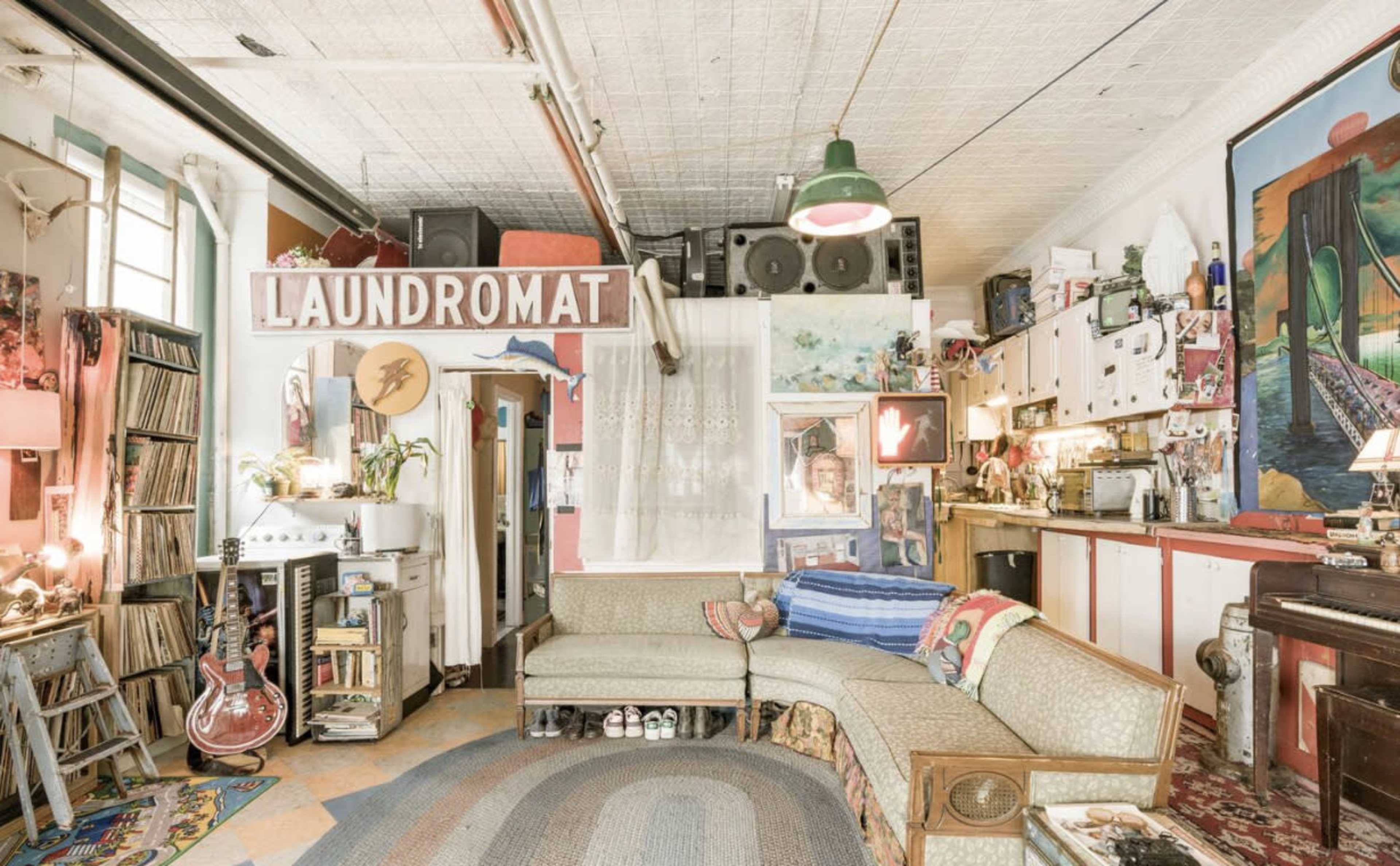 A vibrant and eclectic living space featuring a curved couch, a Laundromat sign, and various pieces of artwork and musical instruments scattered throughout the room.