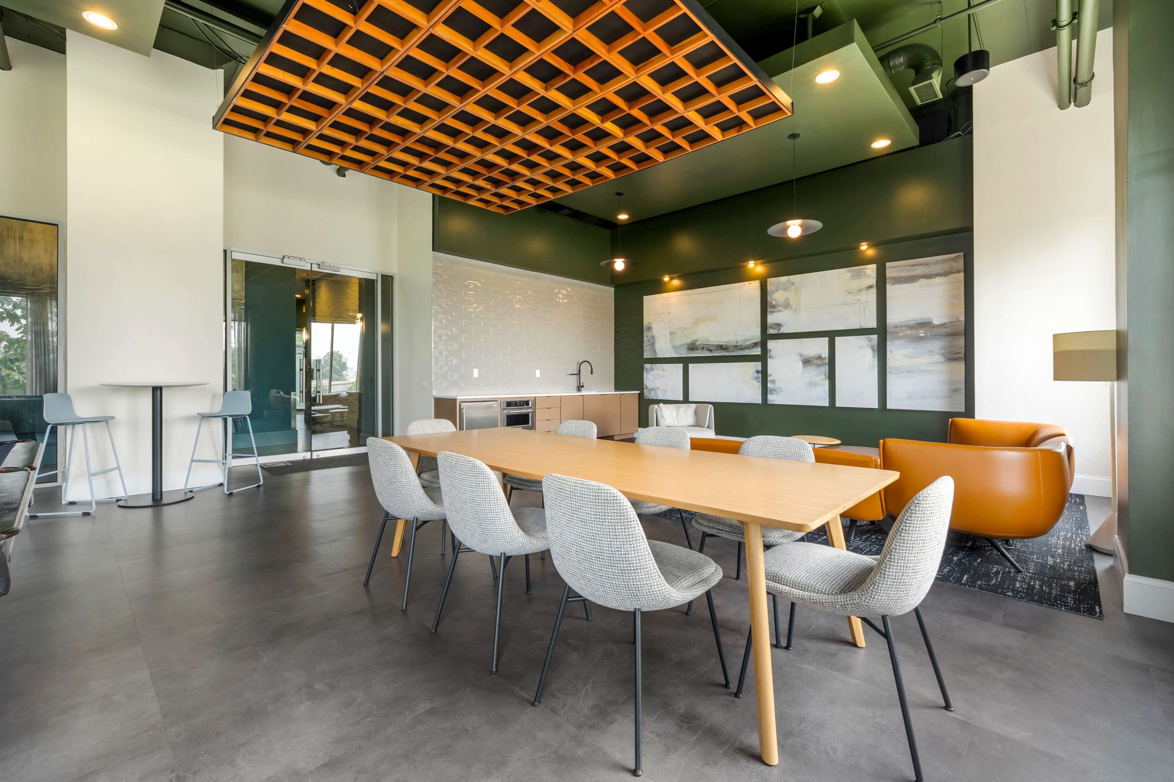 The image shows a modern dining area with a wooden table surrounded by gray chairs, a kitchen in the background, and a lounge area with an orange armchair.