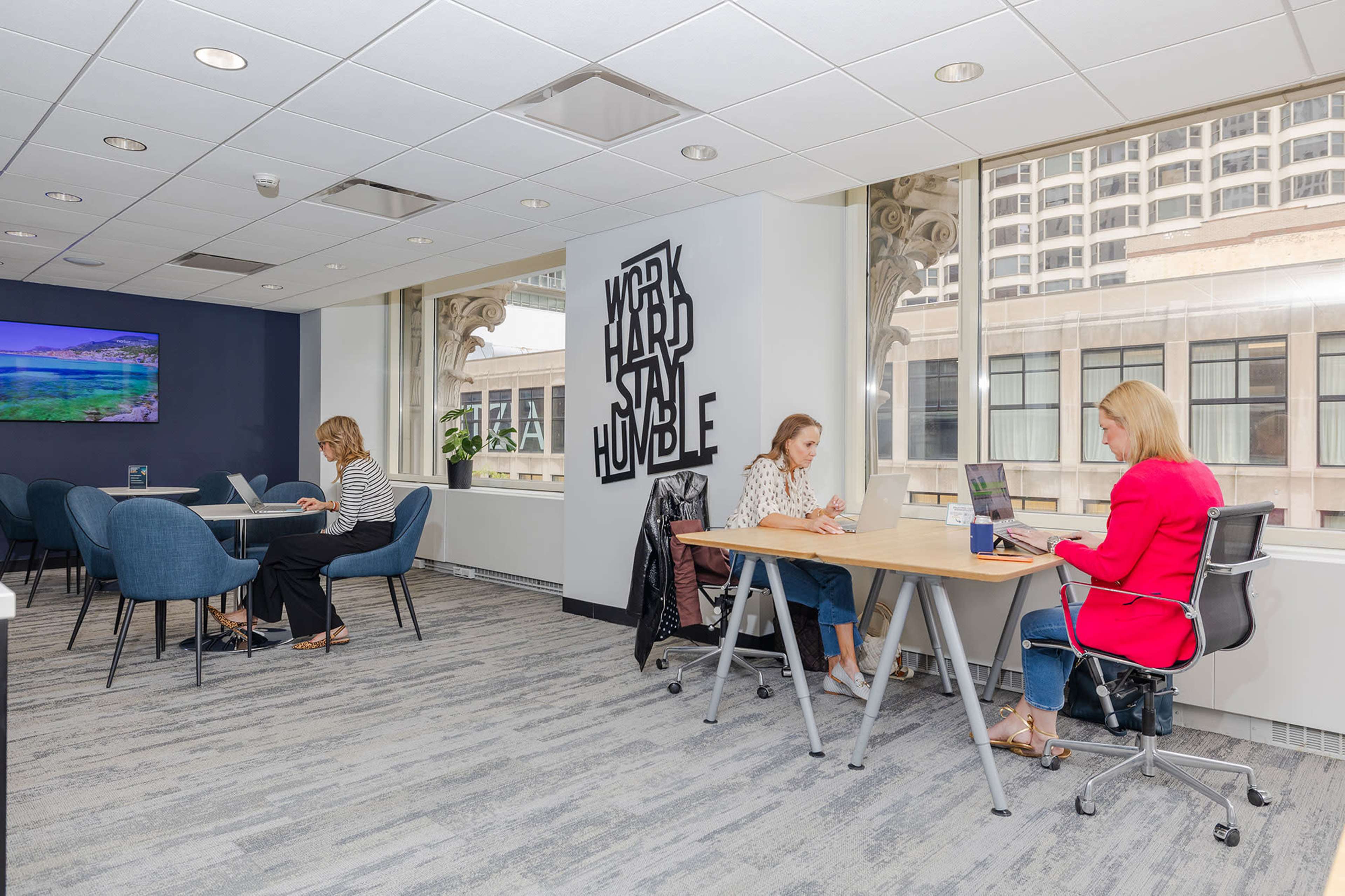 Two women work at desks in a modern office space with large windows and a motivational wall sign that reads "WORK HARD HUMBLE."